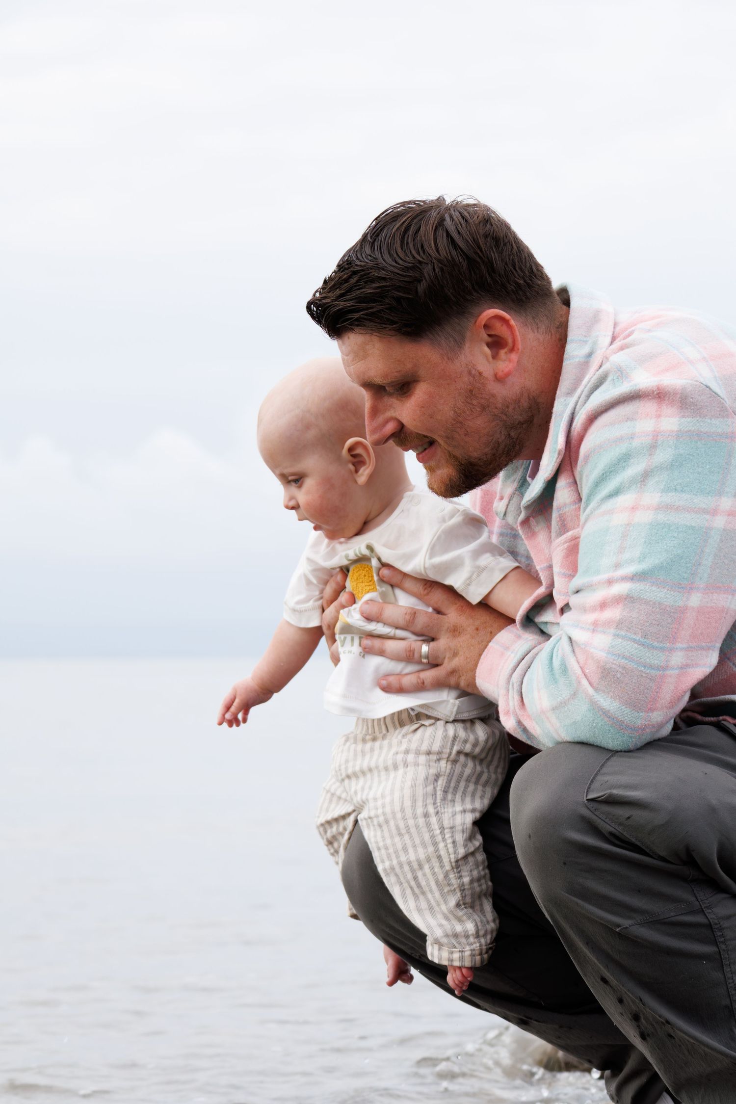 A father crouches by the ocean shore, gently holding his baby as they explore the water together.