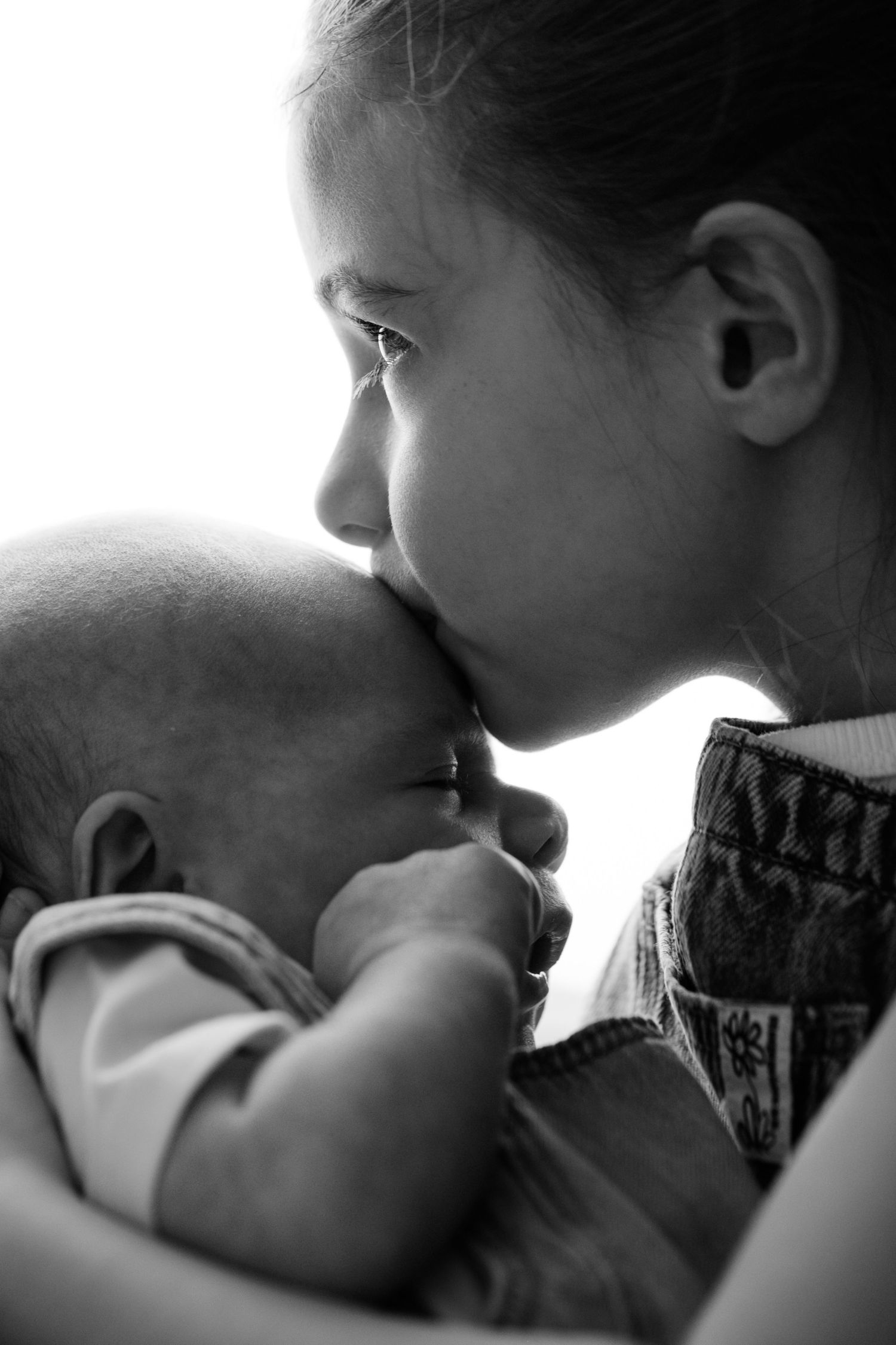 Black and white photo of a young girl tenderly kissing a newborn baby she is holding.