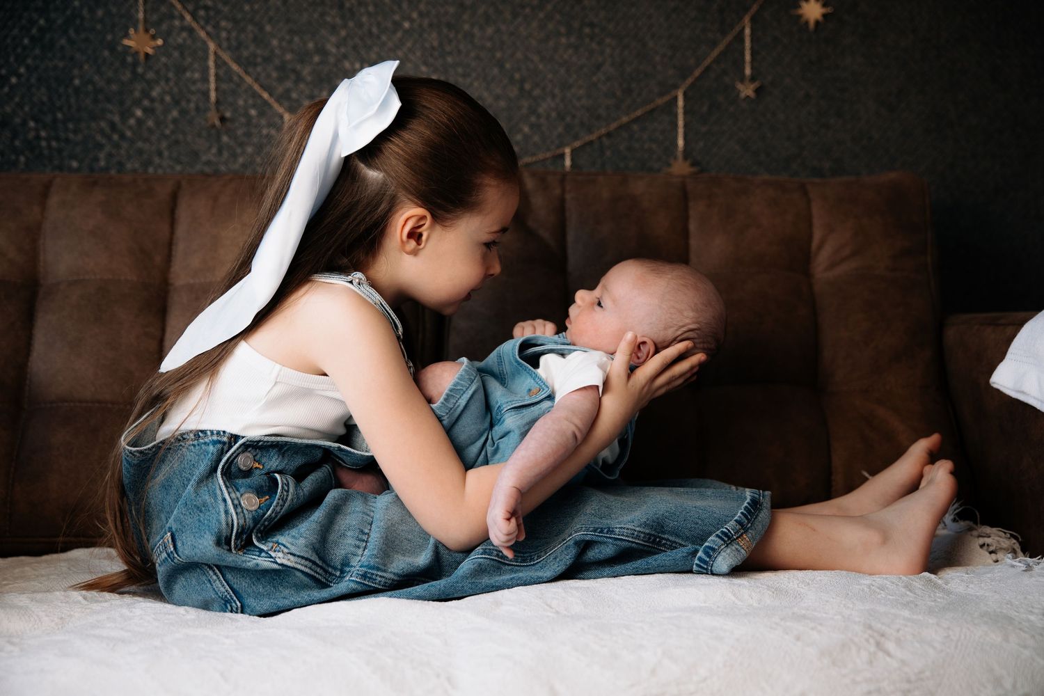 Young girl with white bow gently holds newborn baby sibling on brown sofa in cozy home setting.