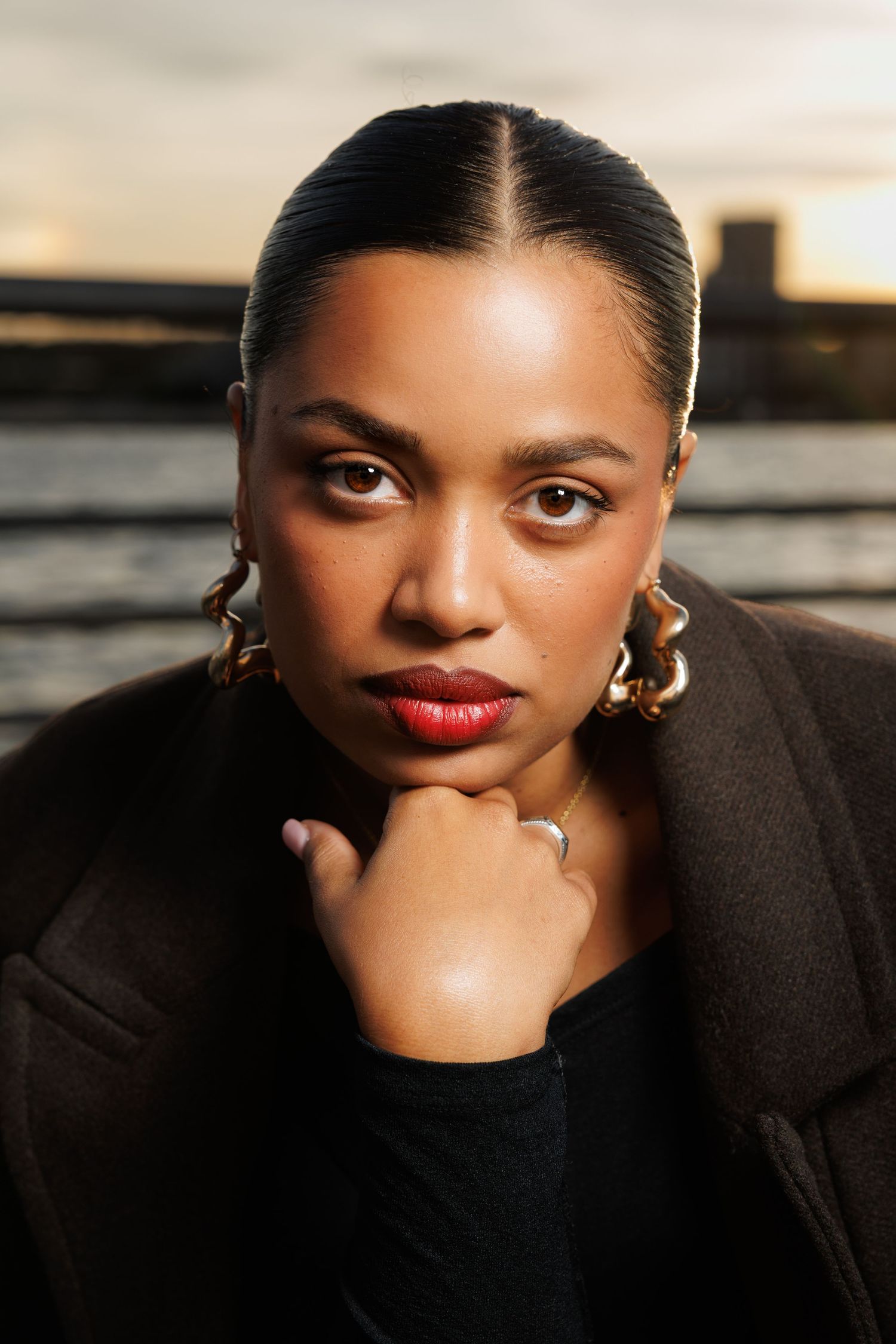 A young Black woman with slicked-back hair, gold hoop earrings, and red lips poses confidently outdoors at sunset.