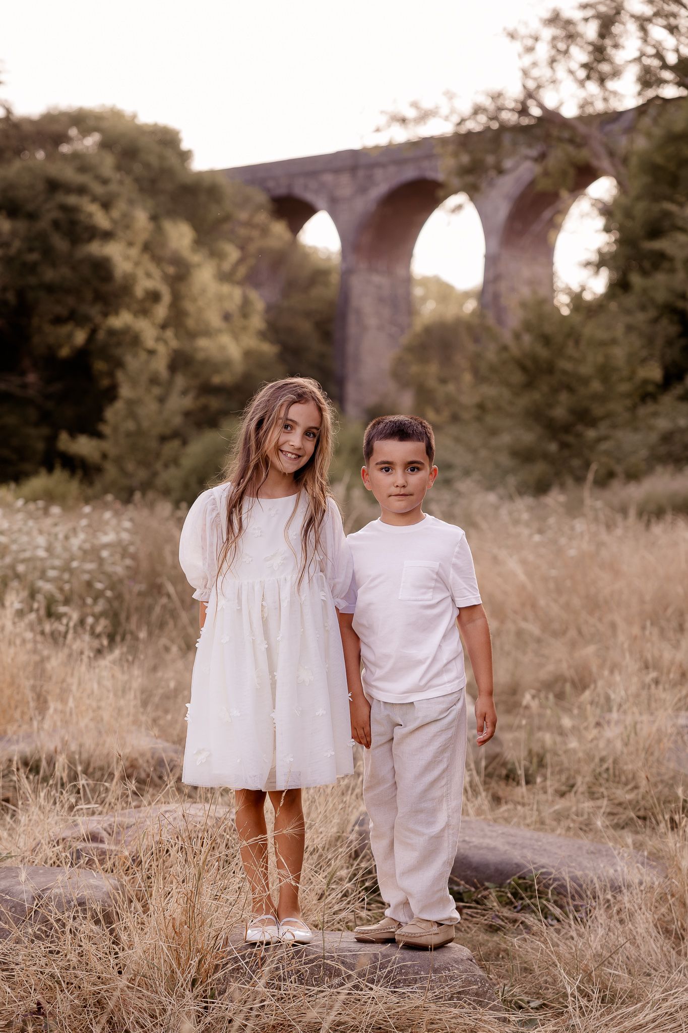 Two children in white outfits stand on rocks in a field with a stone arch viaduct in the background.