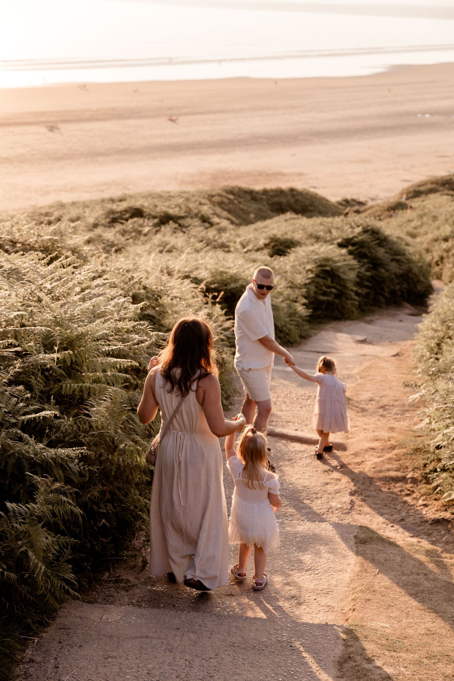 Family of four walking on a sandy beach path at golden hour, surrounded by lush green dunes near the ocean.