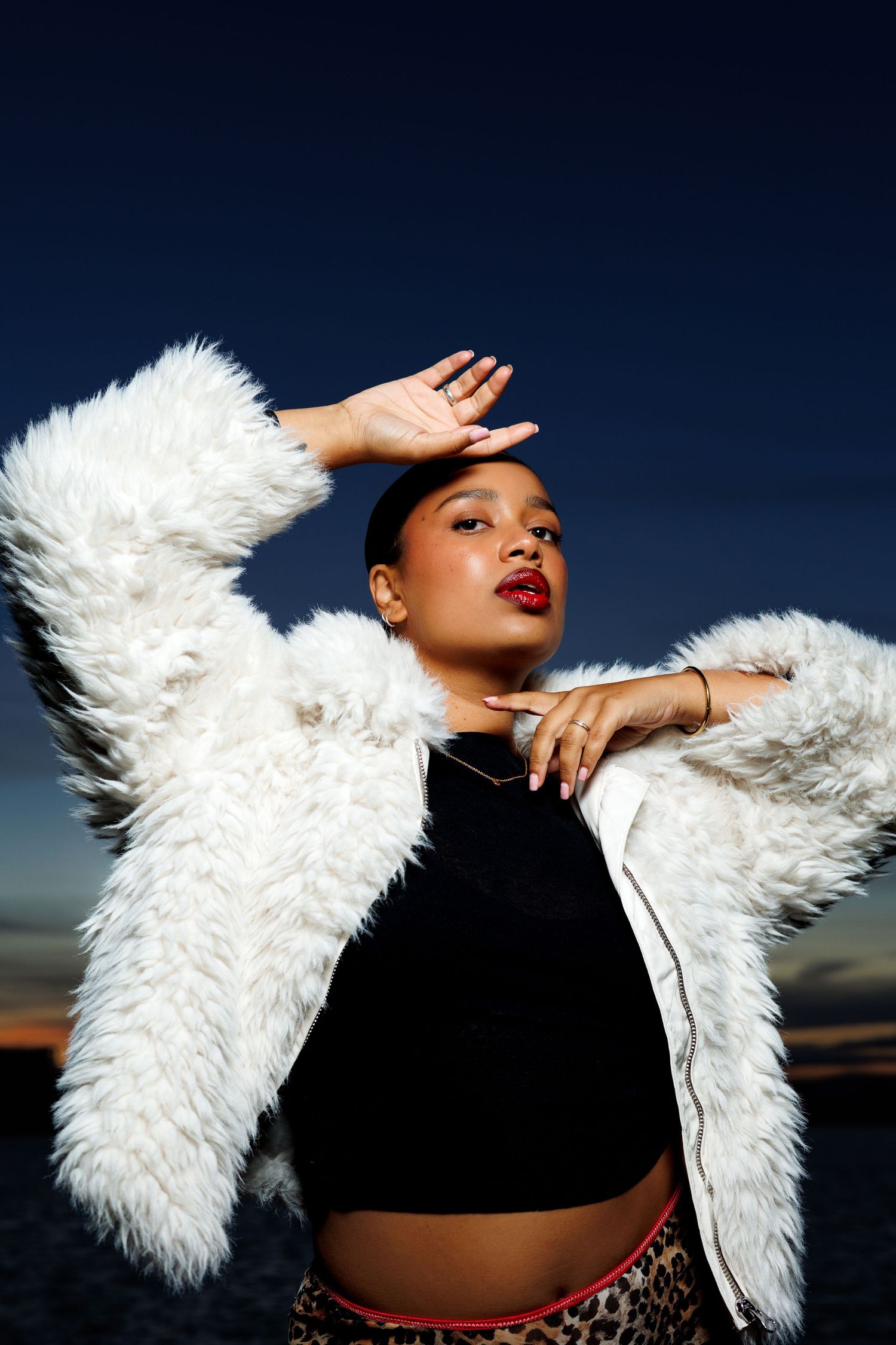 Woman in white faux fur jacket and crop top poses against twilight sky near water with red lips.