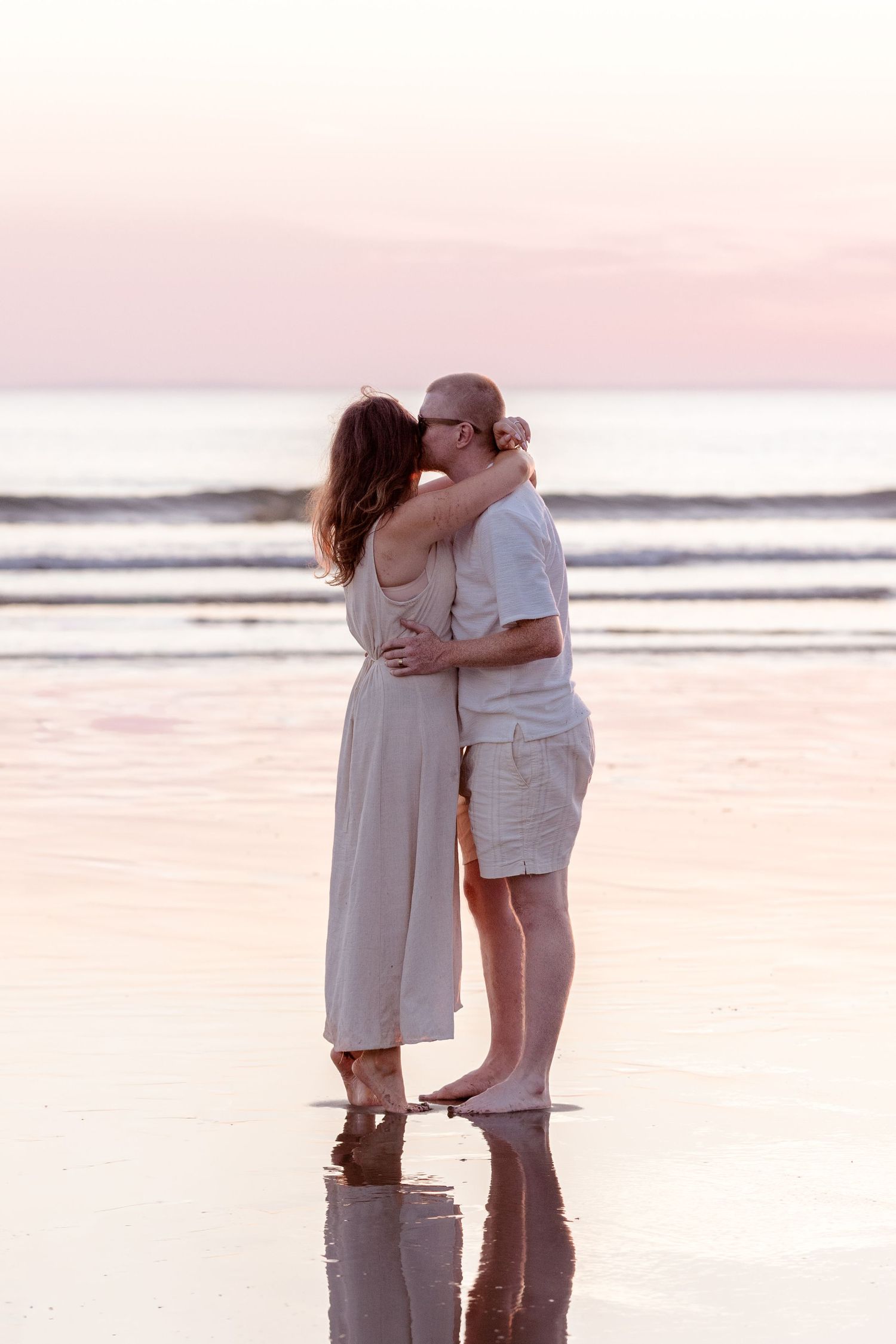 Couple sharing a romantic kiss on a reflective beach at sunset, dressed in light casual clothing.