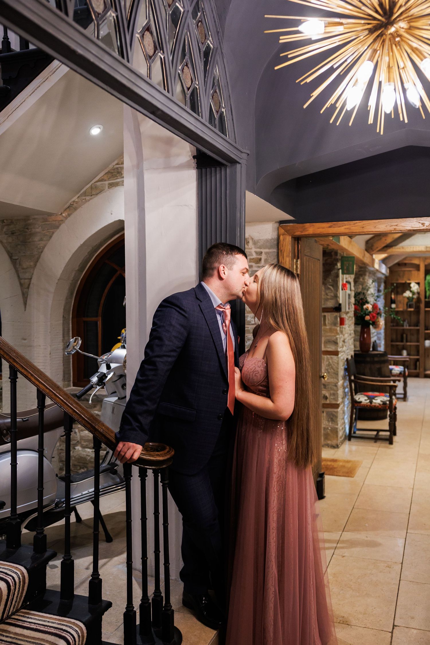 Couple sharing a romantic kiss on a staircase in an elegant venue with a gold sputnik chandelier overhead.