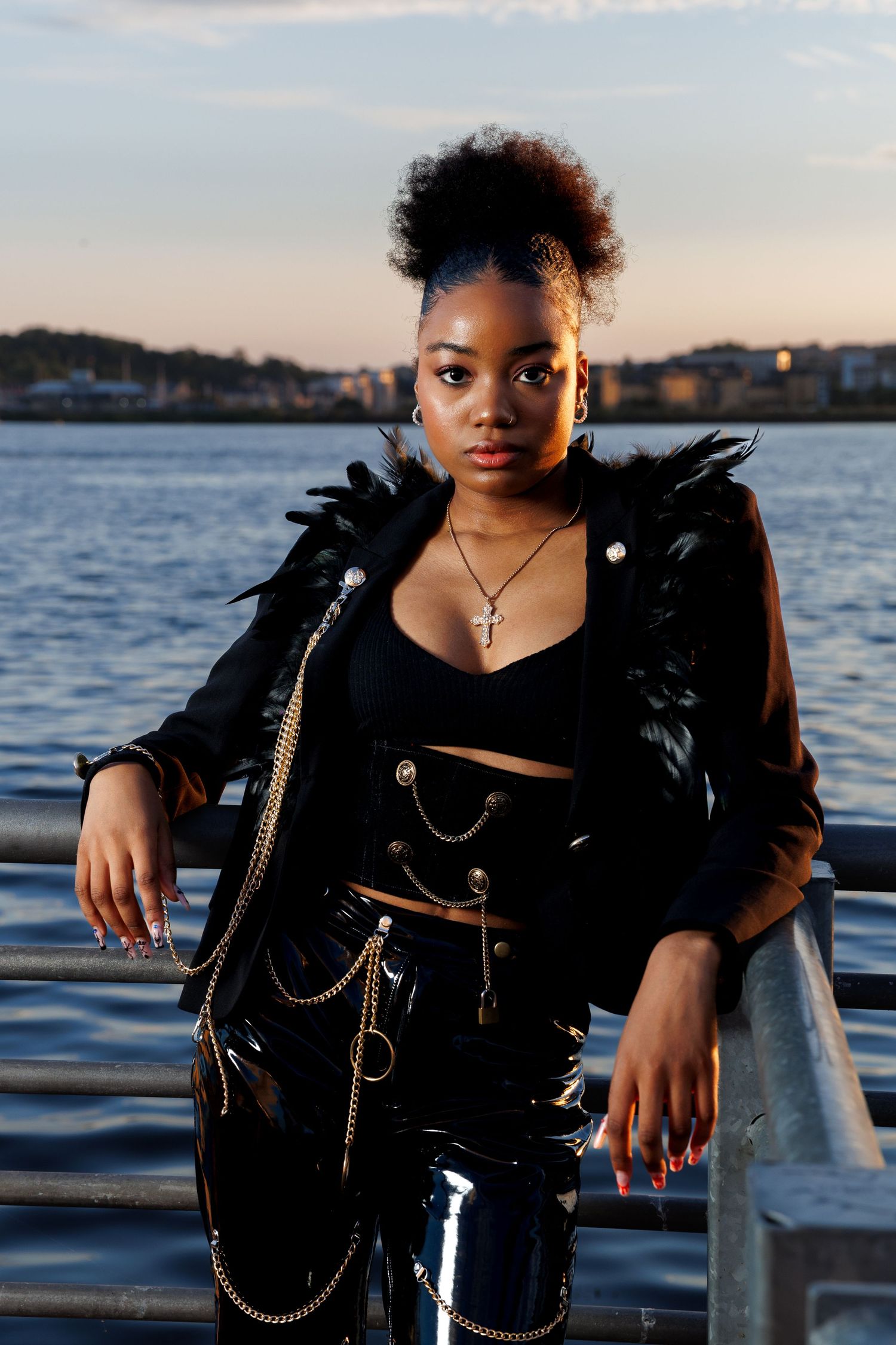 Young Black woman in black feathered jacket, vinyl pants, and gold chains poses by waterfront railing at dusk.