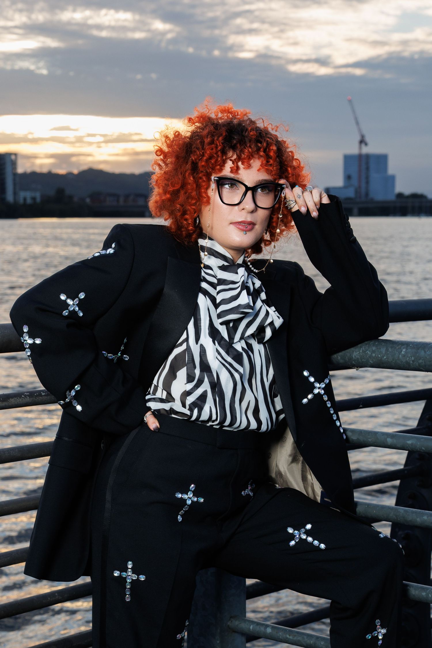Curly red-haired woman in black cross-print suit and glasses posing by waterfront at dusk.