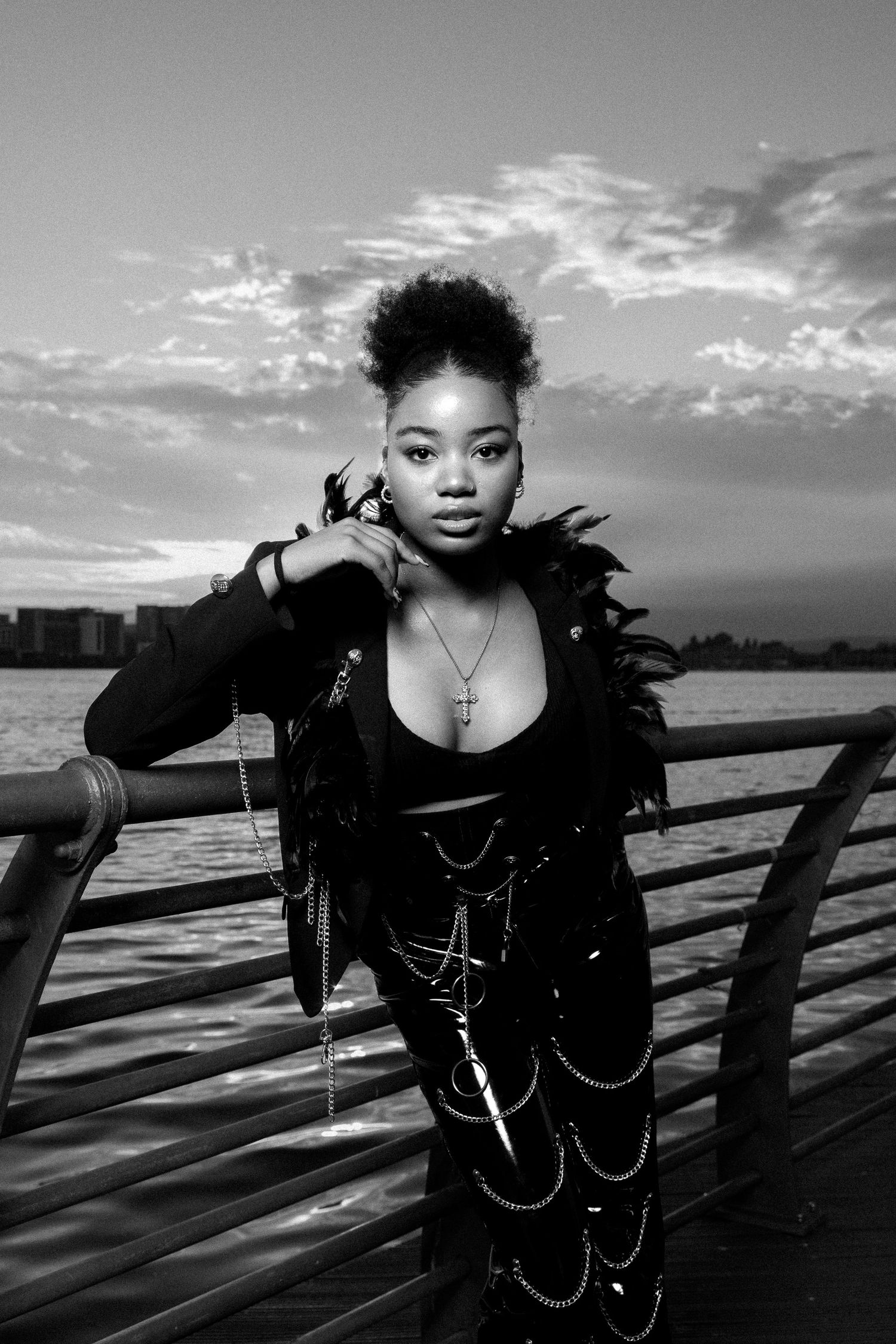 Black and white photo of a young woman in edgy chain outfit with feather boa posing on a waterfront pier at dusk.