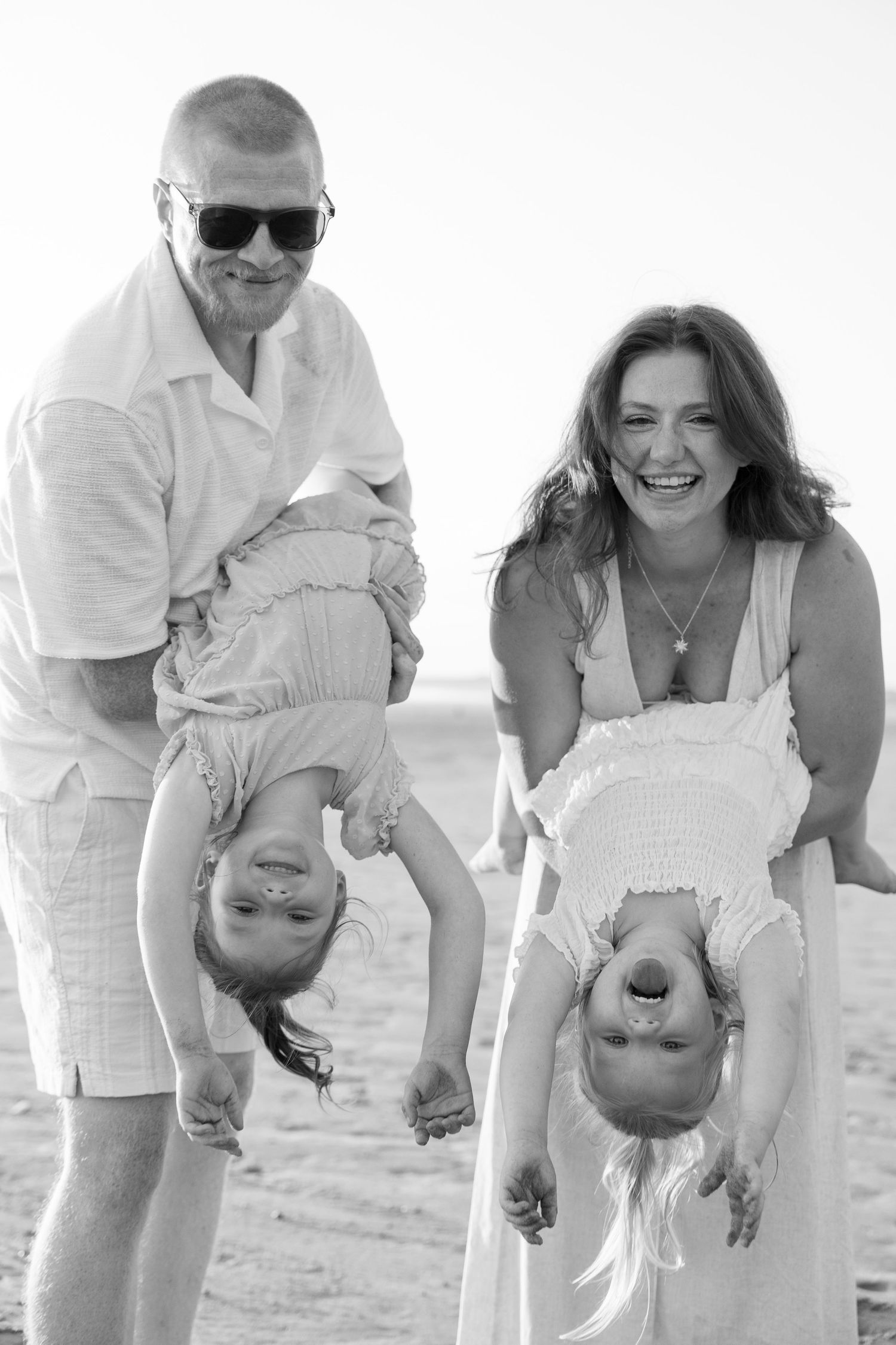 Black and white photo of smiling parents holding two young children upside down on a sunny beach.