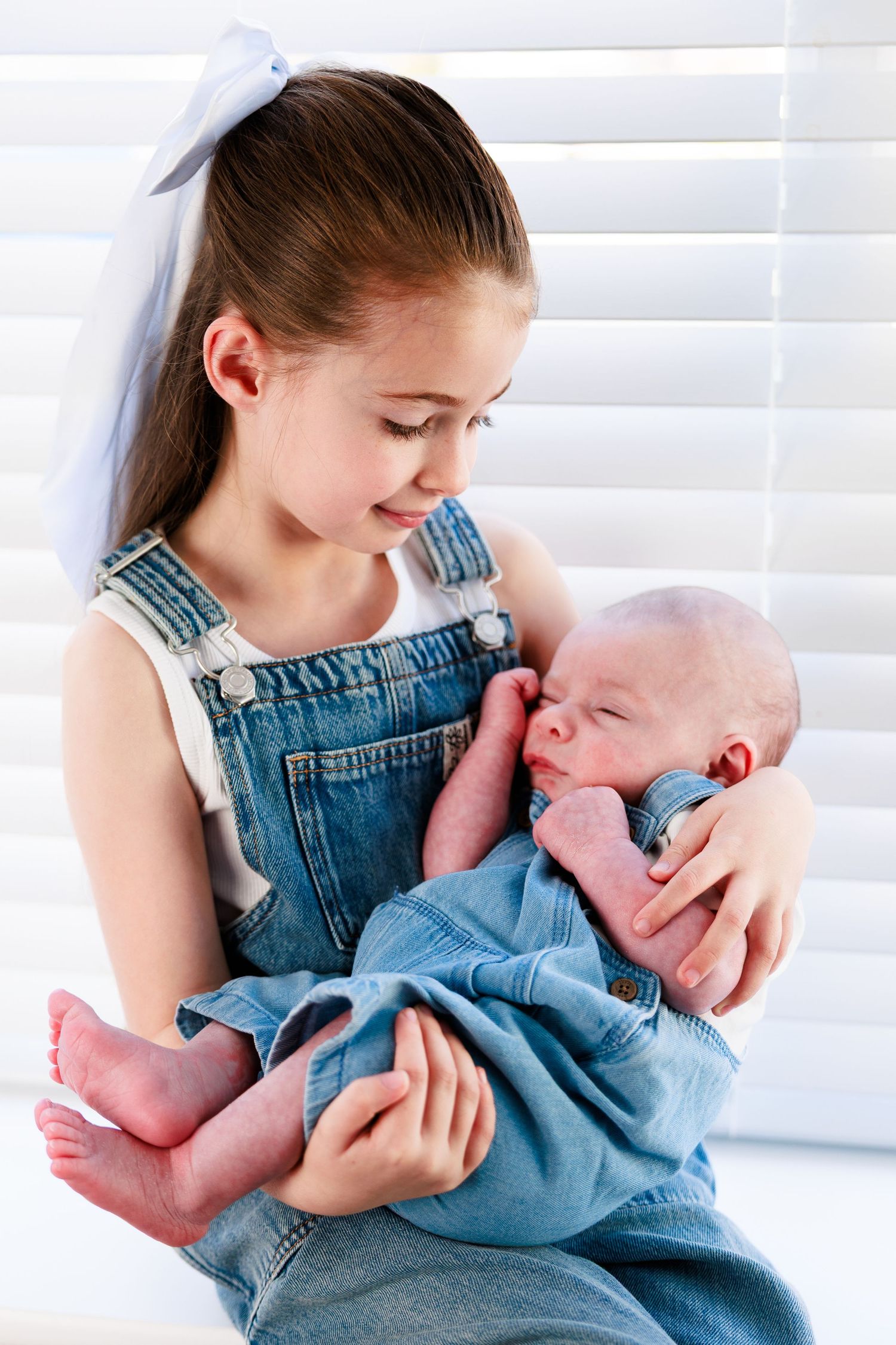 Young girl in denim overalls carefully holds a sleeping newborn baby near window blinds.