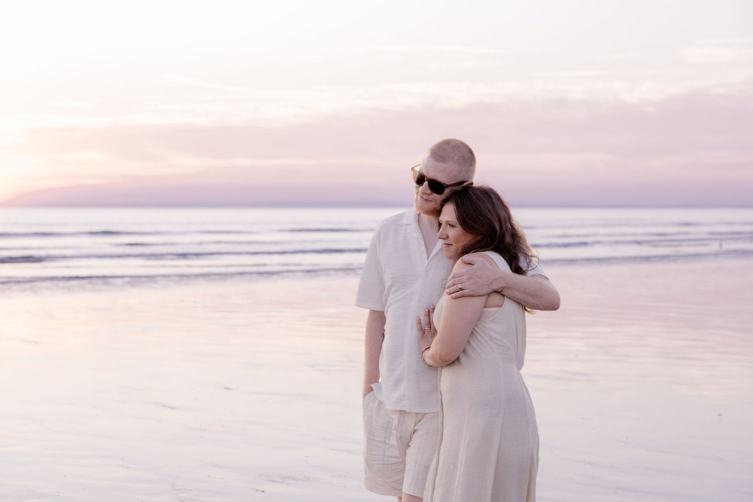 Couple embracing tenderly on a beach at sunset with pastel pink sky reflecting on water.