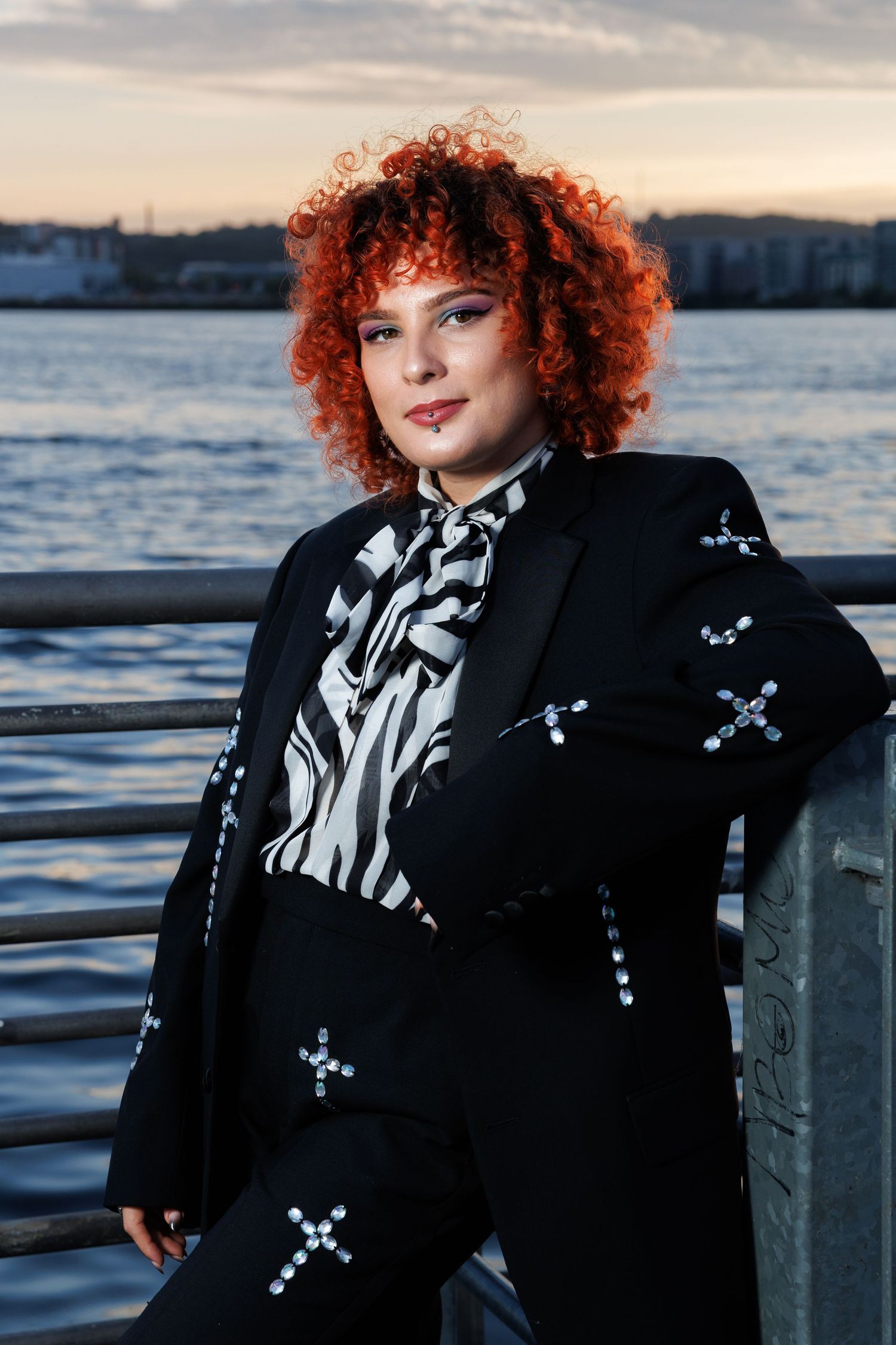Woman with red curly hair in black cross-embroidered suit and zebra print blouse poses at waterfront sunset.
