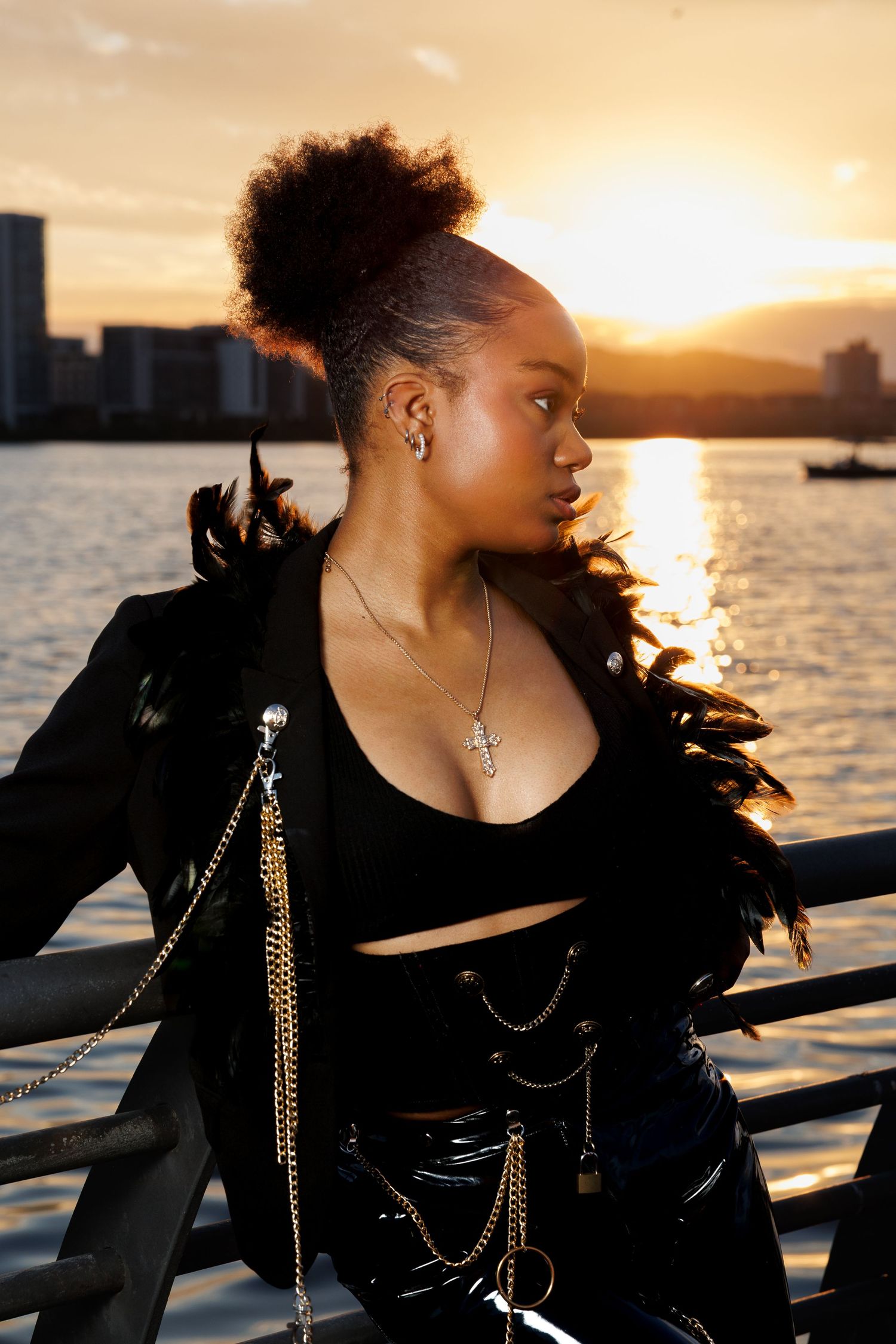 Black woman in edgy feather jacket and chains posing by waterfront railing at golden sunset.