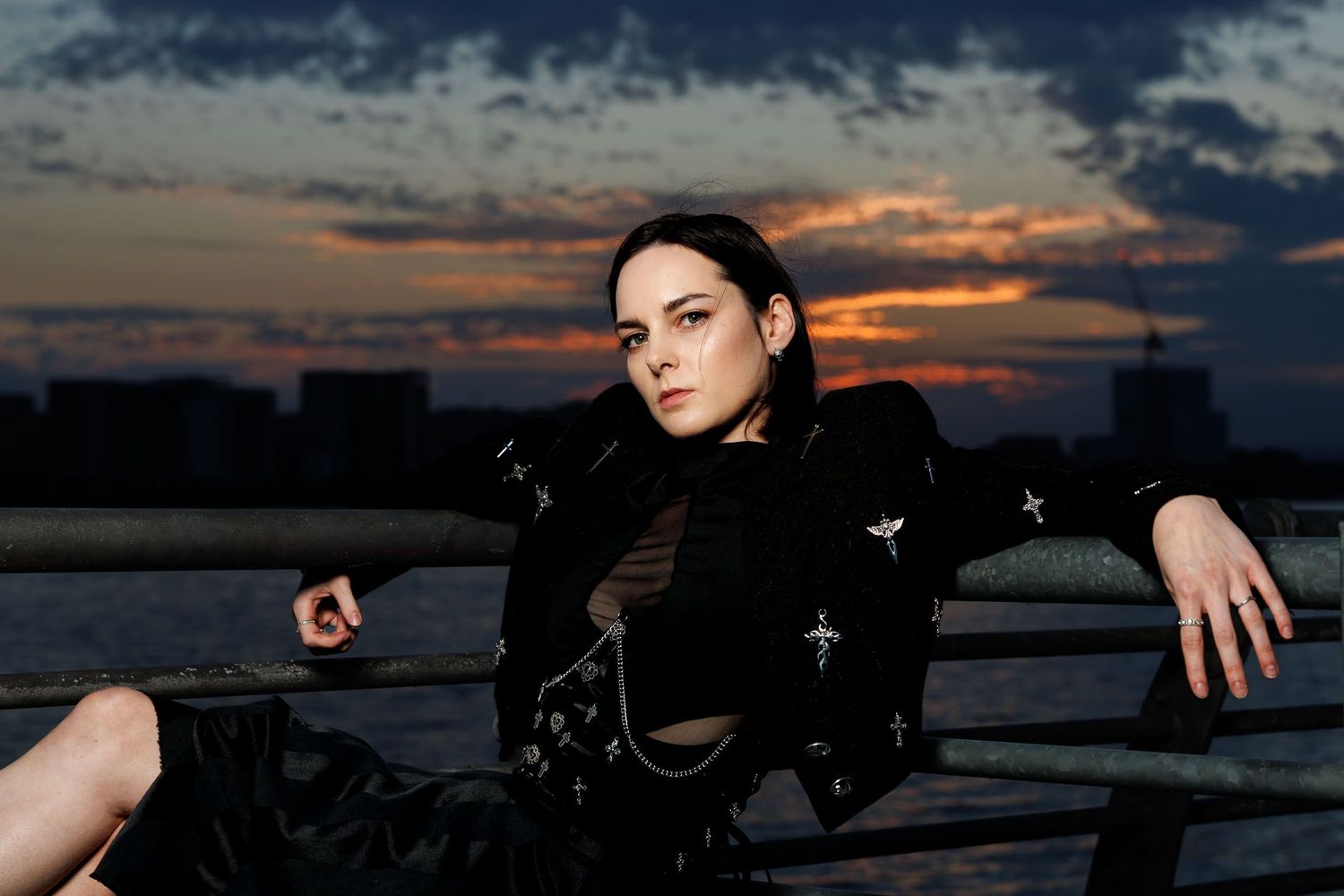A young woman in dark clothing poses on a railing at sunset with a dramatic sky and city skyline behind her.