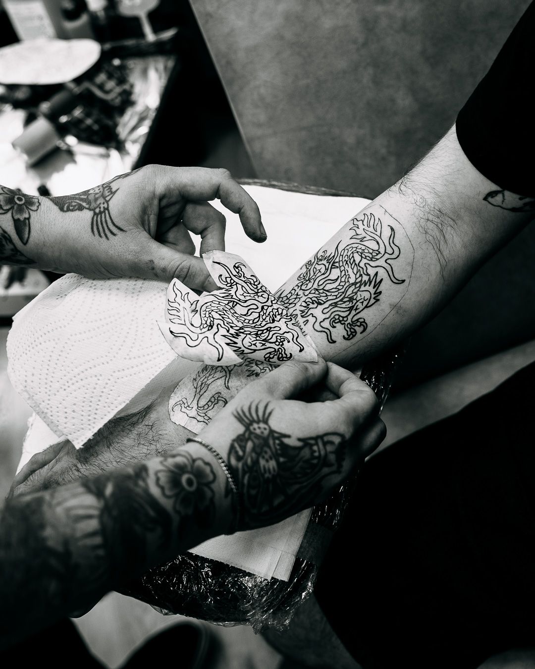 Black and white close-up of a tattoo artist applying a detailed dragon tattoo stencil to an arm.