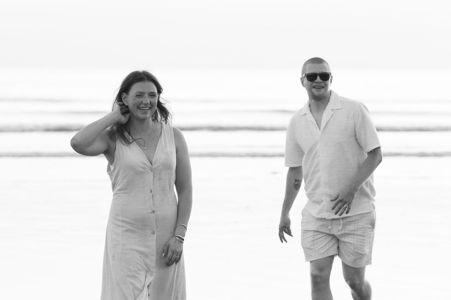 Couple walking on beach in black and white photo, woman in sundress and man in casual shirt with sunglasses.