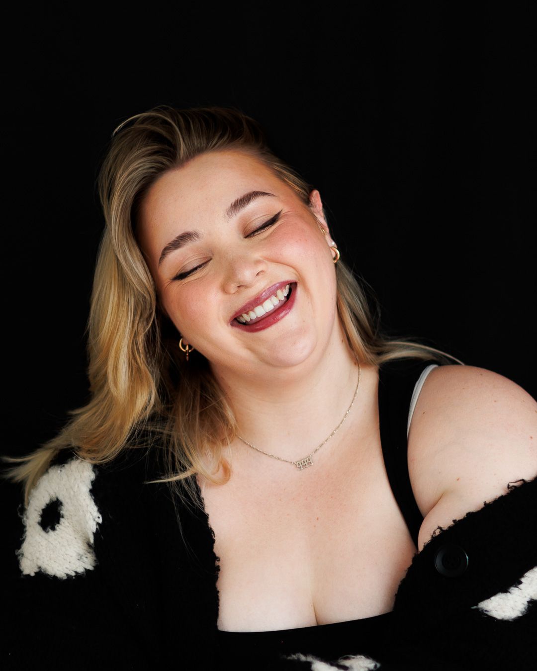 Laughing blonde woman in a black and white outfit poses against a dark background.