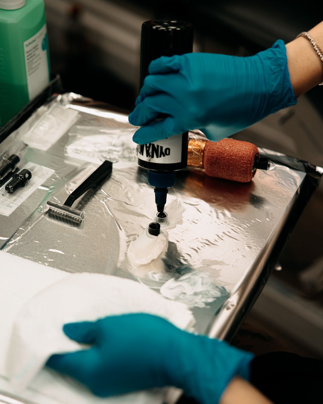 Tattoo artist in blue gloves filling ink into tattoo machine on foil-covered tray.