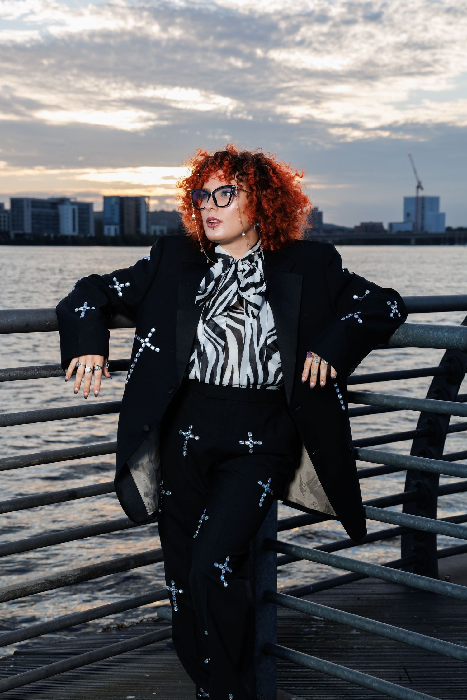 Curly red-haired woman in black cross-embroidered suit and zebra print blouse poses at waterfront pier.