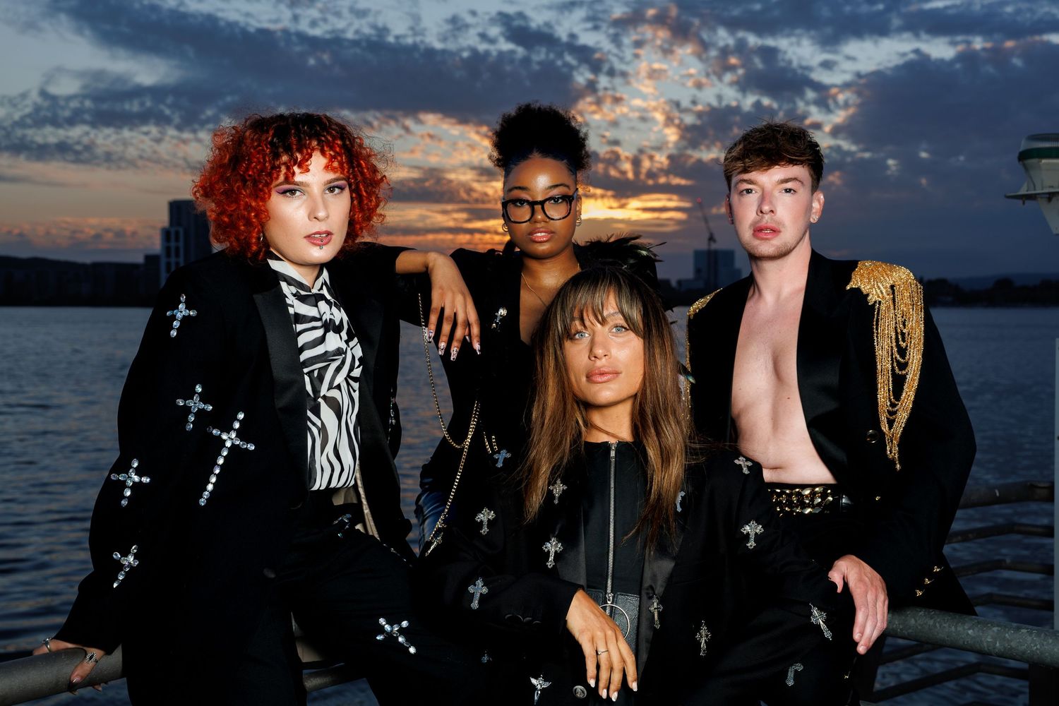 Four stylish young people pose together at sunset by the water, wearing black outfits with cross details.