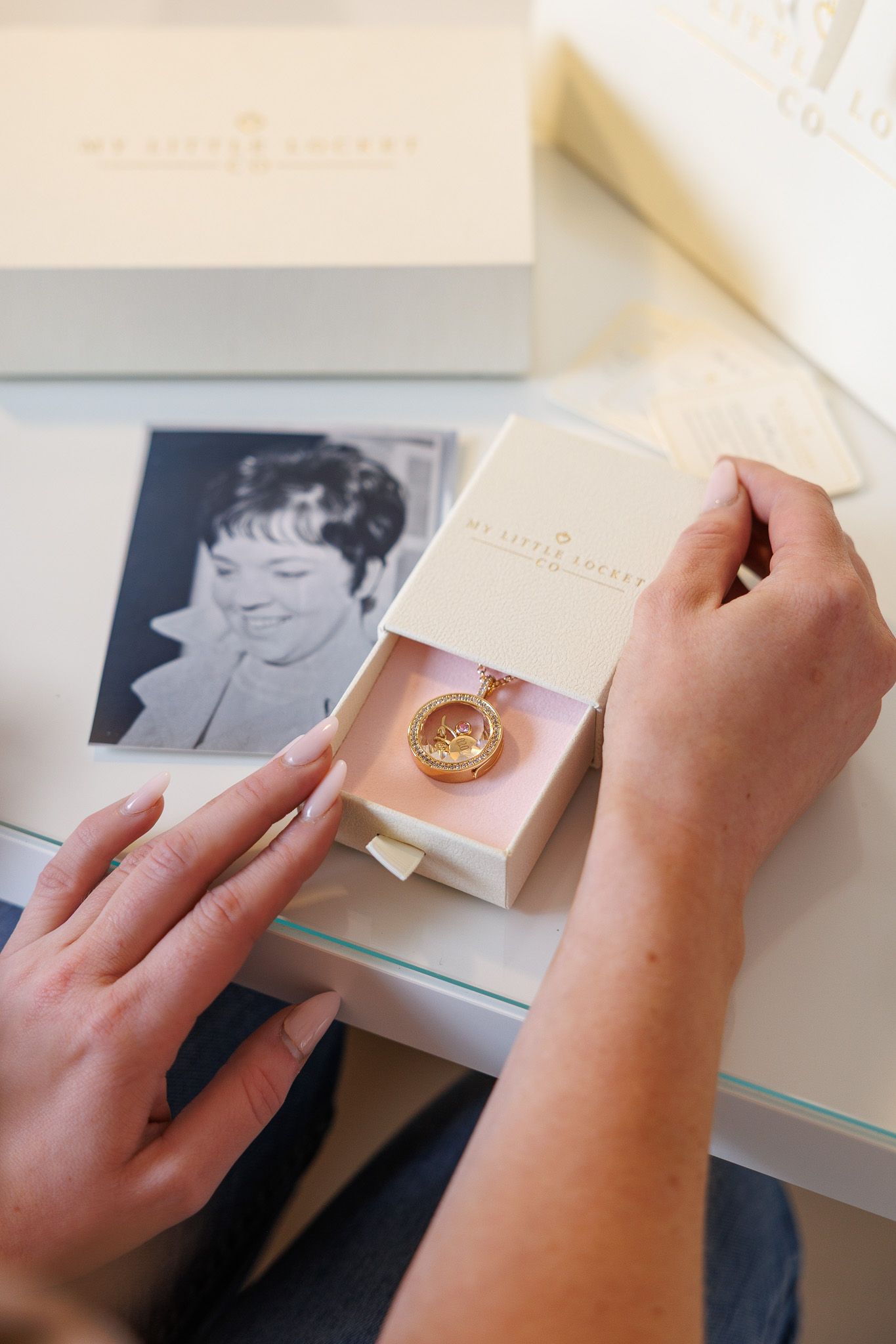 Hands opening a luxury gift box containing a gold locket necklace, with a vintage black and white photo in the background.