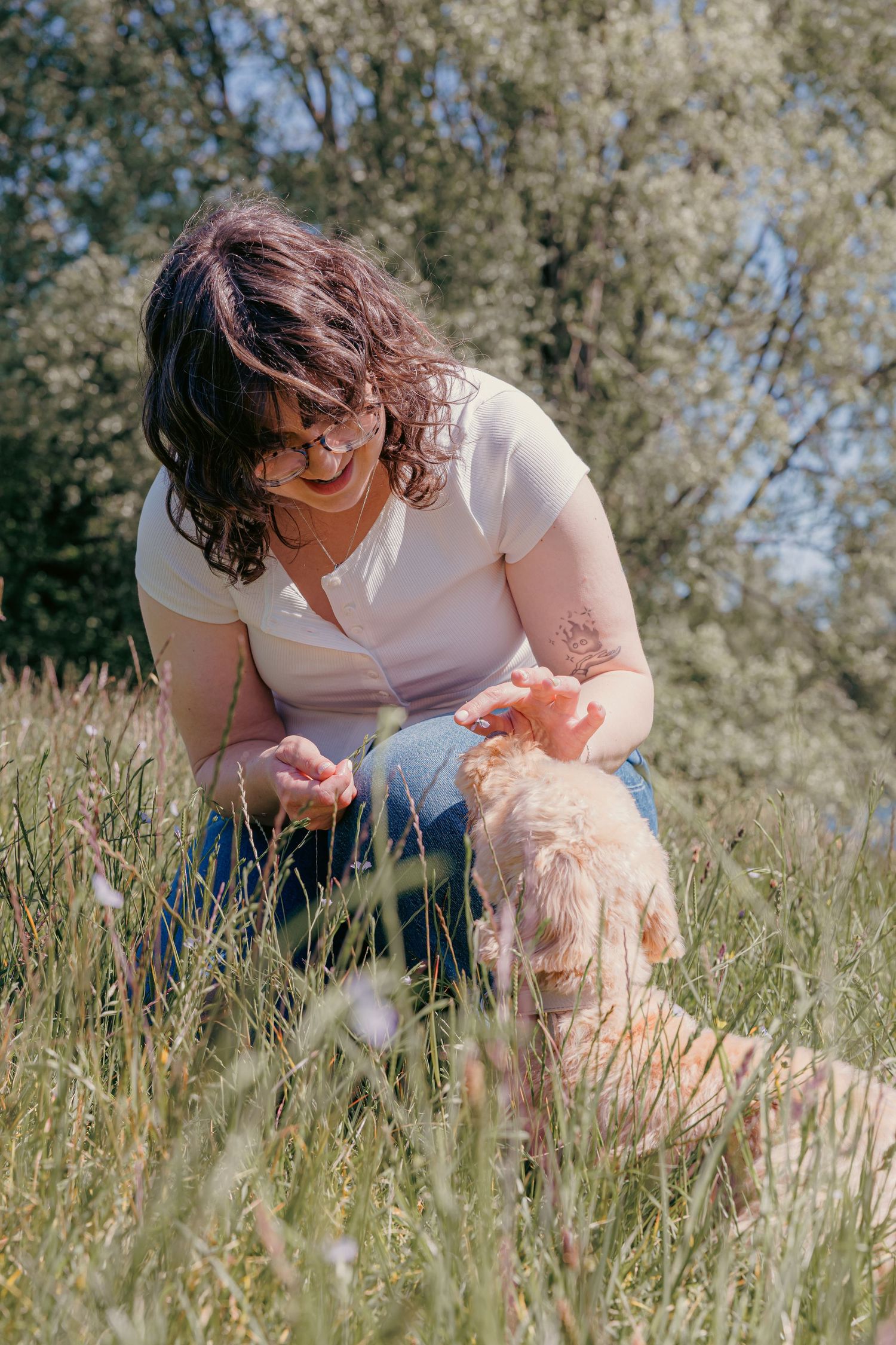 Woman in white top and jeans kneeling in tall grass, petting a golden dog outdoors on a sunny day.