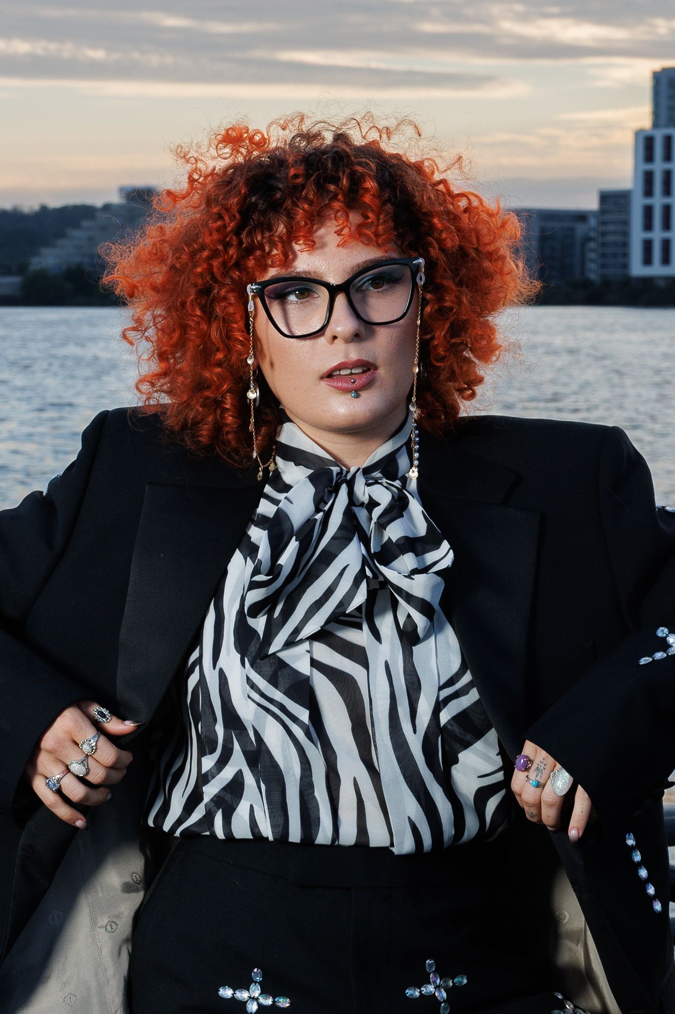 Curly red-haired woman in glasses and zebra-print blouse with black blazer posing near urban waterfront at dusk.