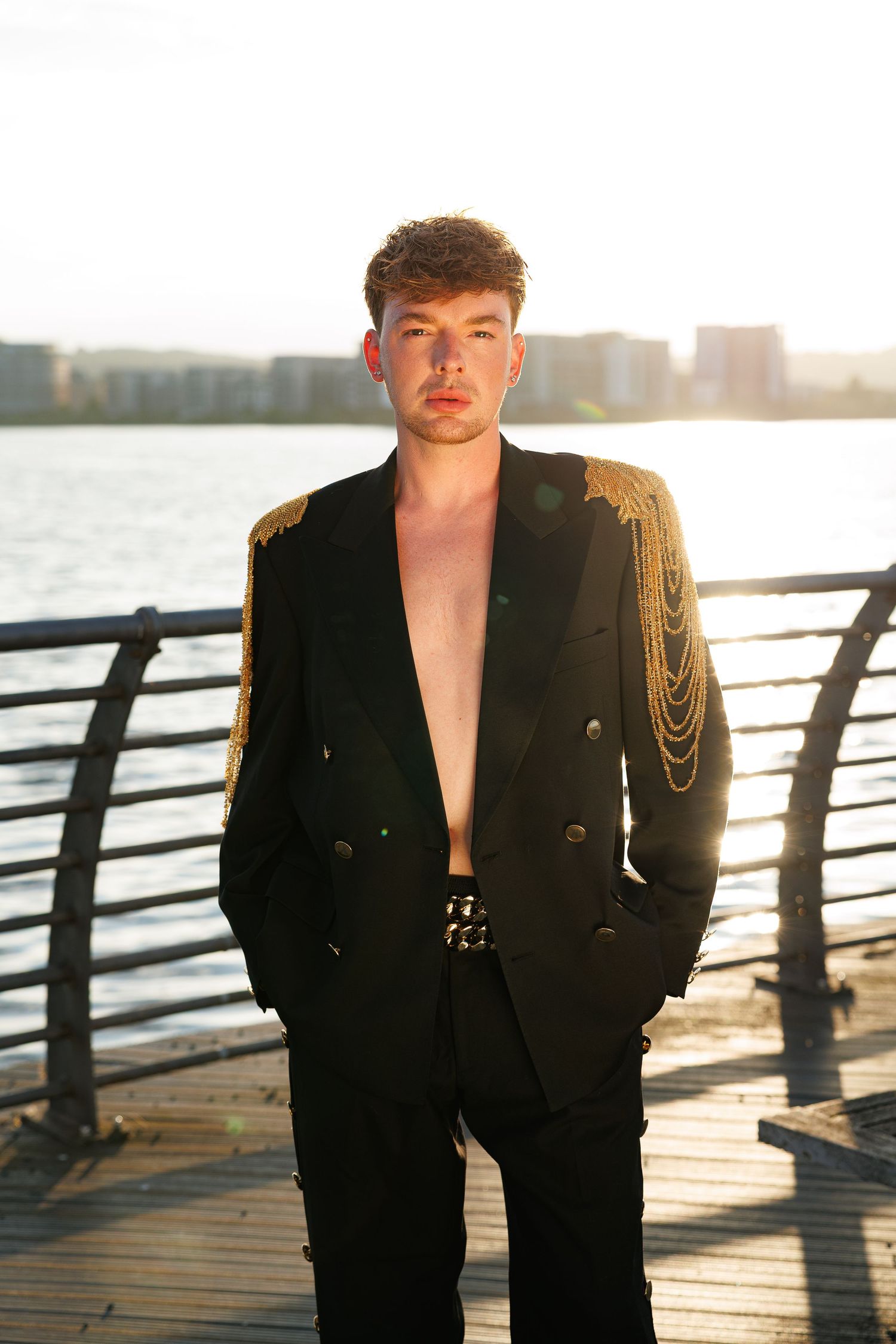 Young man in black military-style blazer with gold epaulettes poses shirtless on waterfront pier at golden hour.