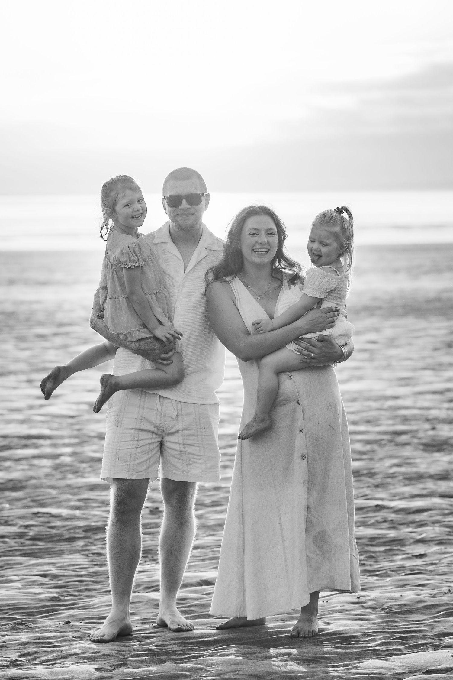 Black and white photo of a happy family of four posing on a beach at sunset, parents holding two young daughters.
