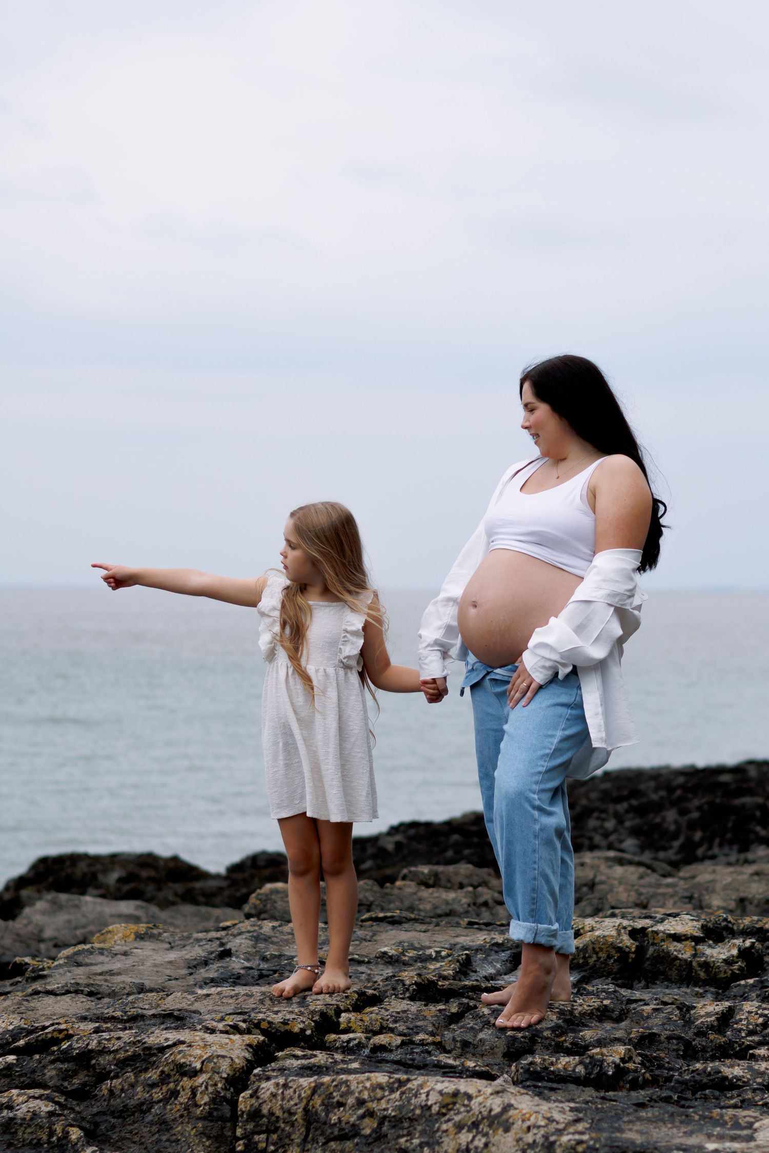 Pregnant woman in white top and jeans holds hands with young girl on rocky beach, both barefoot by the ocean.