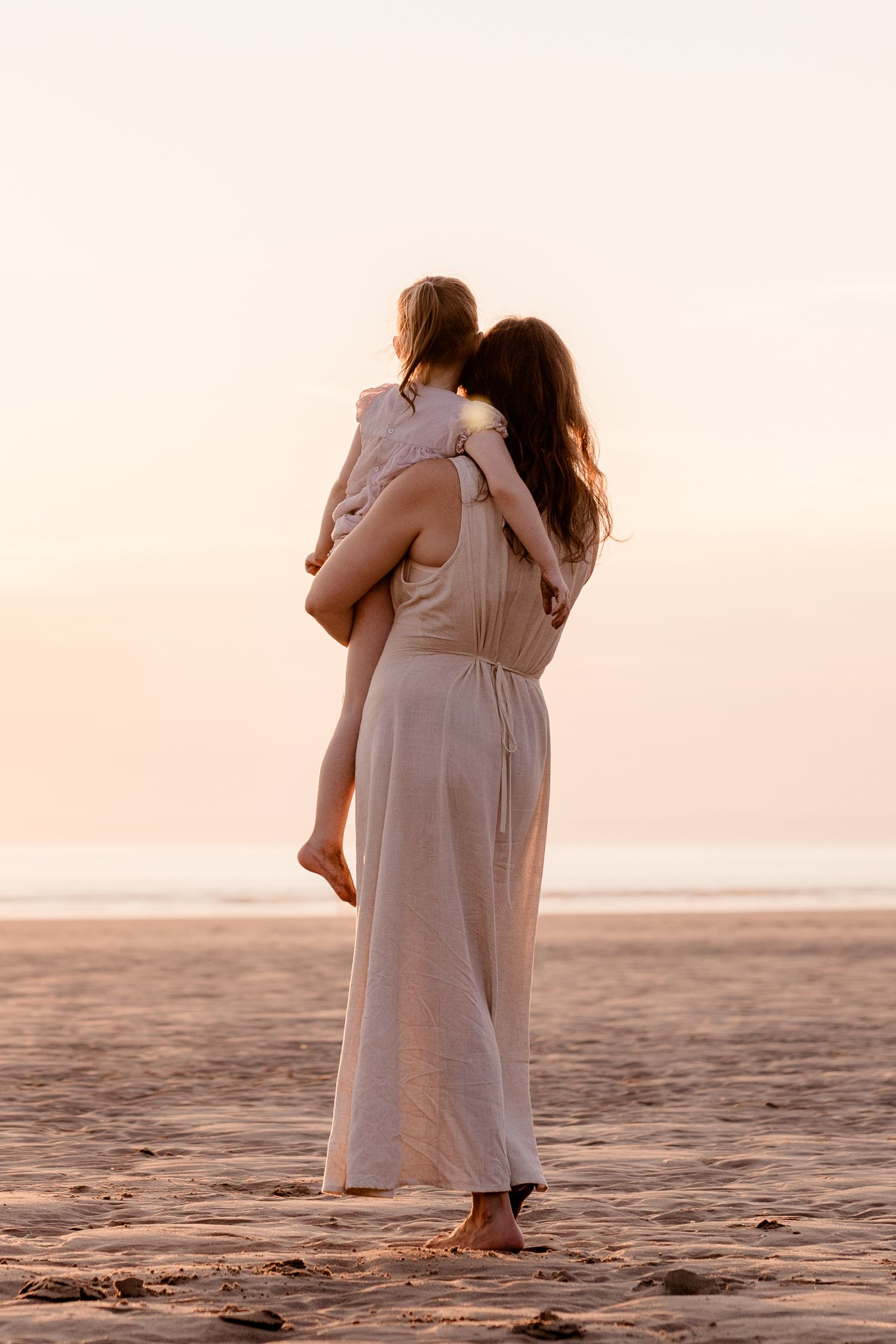Mother in white dress holds child on beach at sunset, standing barefoot on sand with ocean in background.