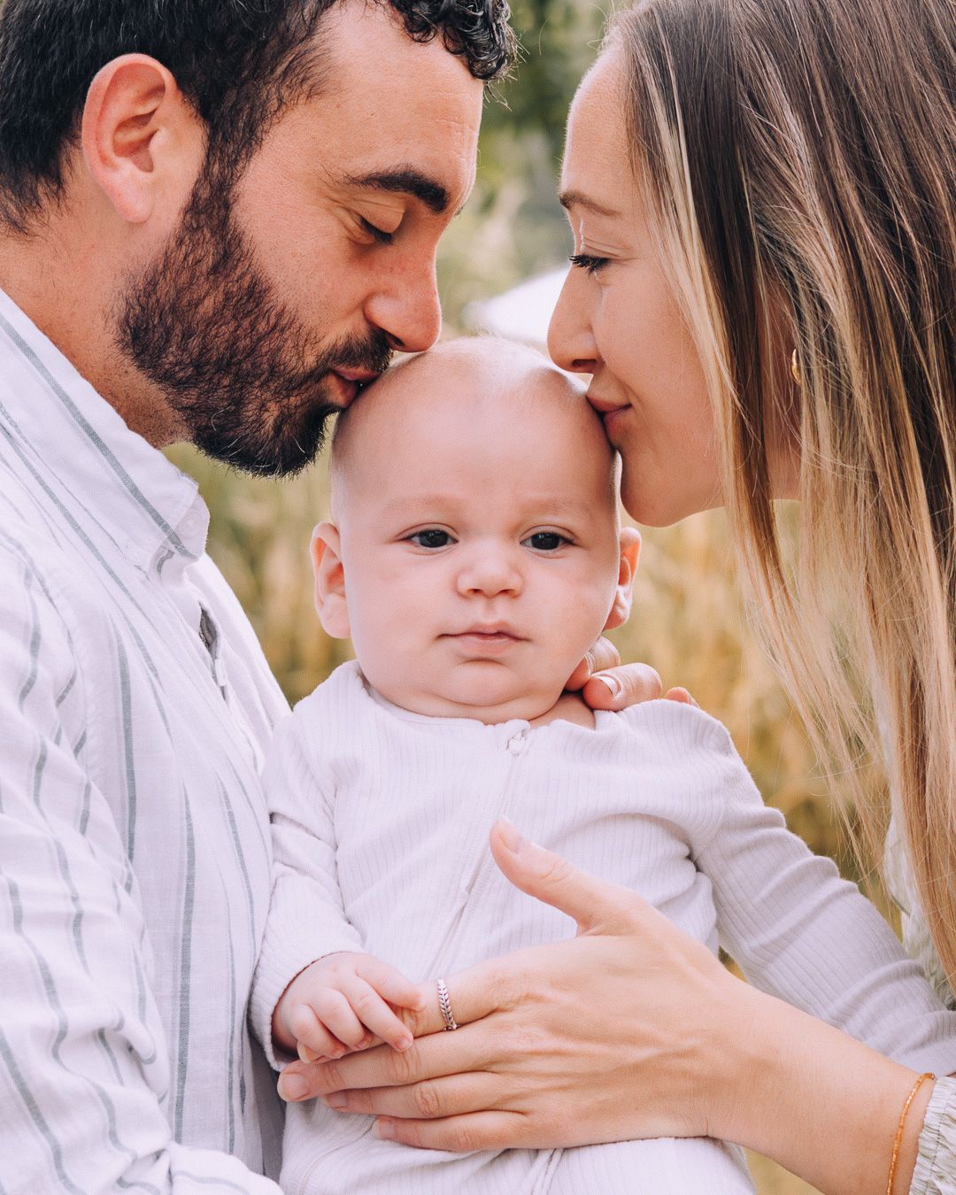 A couple lovingly kisses their adorable baby on the cheeks outdoors in a tender family portrait.