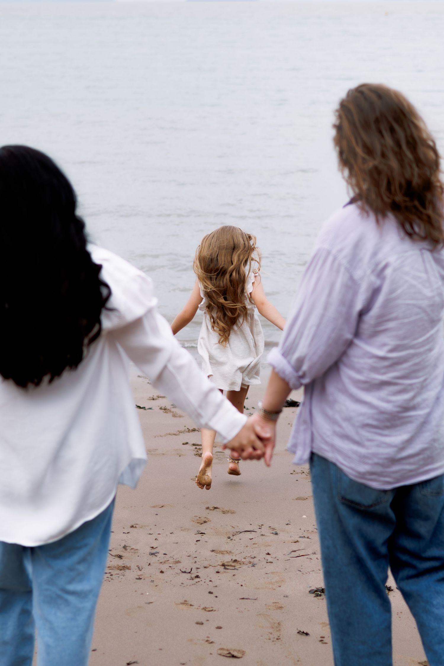 Family walking on beach holding hands, parents and child in white dress running toward calm water.