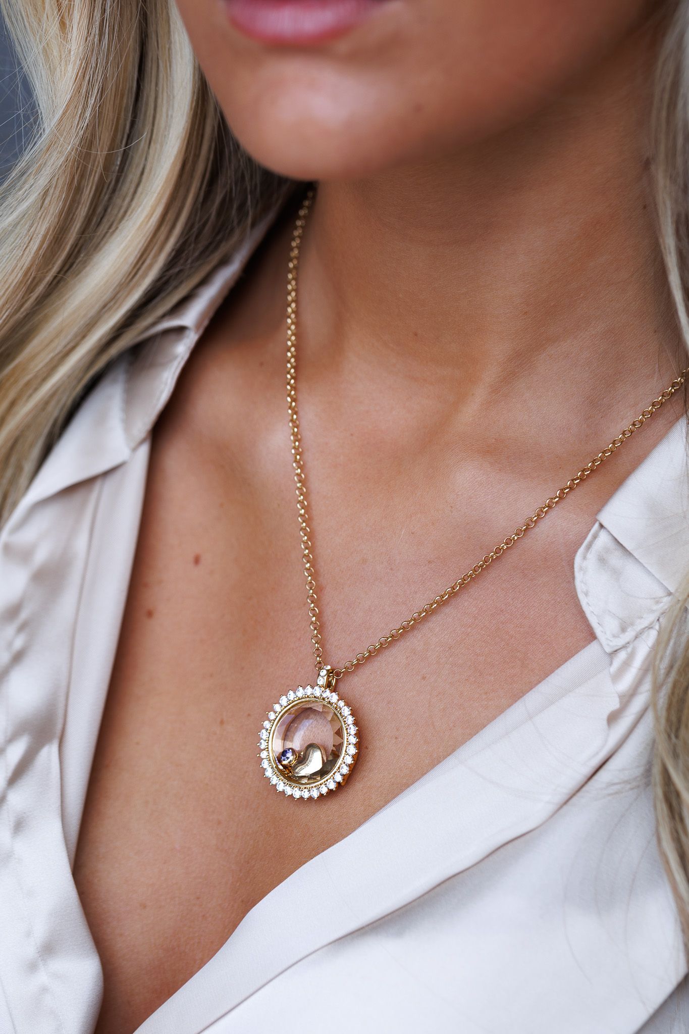 Gold chain necklace with crystal-rimmed circular pendant on a blonde woman wearing a satin blouse.