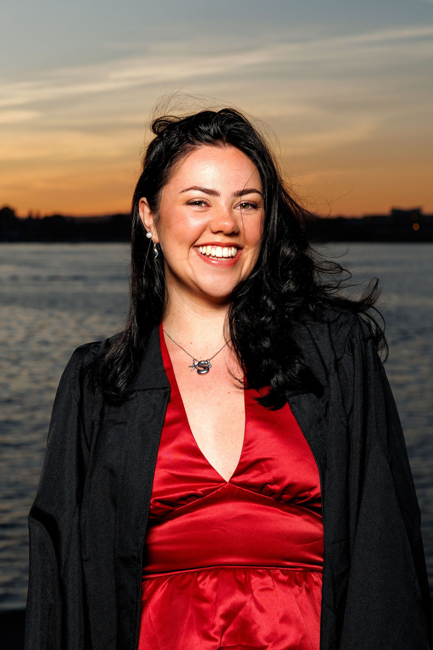 Smiling woman in red satin dress and black graduation gown poses by waterfront at sunset.