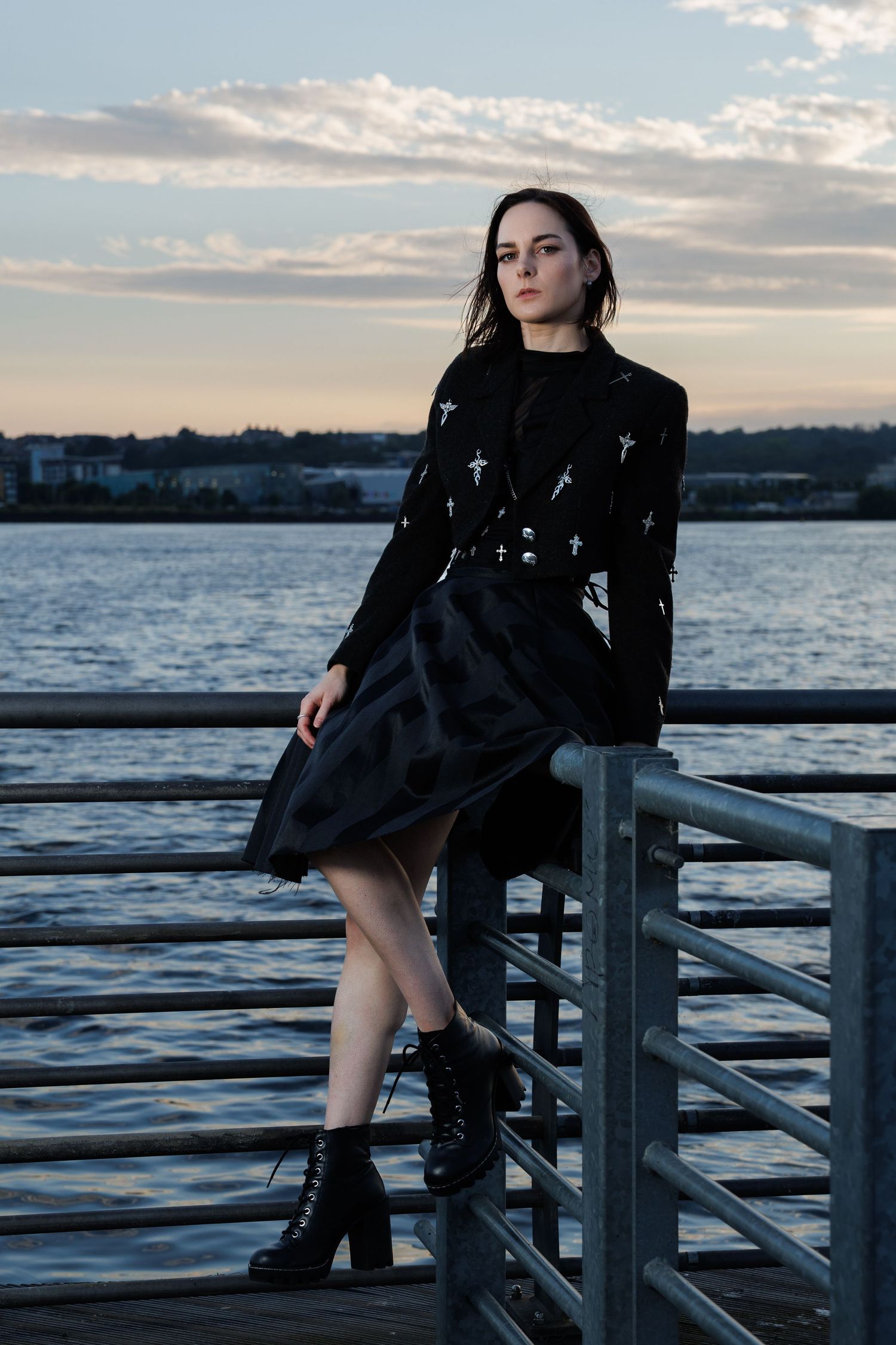 Woman in black cross-embroidered blazer and skirt leans on metal railing at waterfront during sunset.