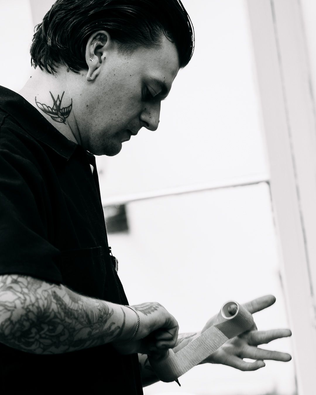 Black and white portrait of tattooed artist with neck tattoo examining his hand.