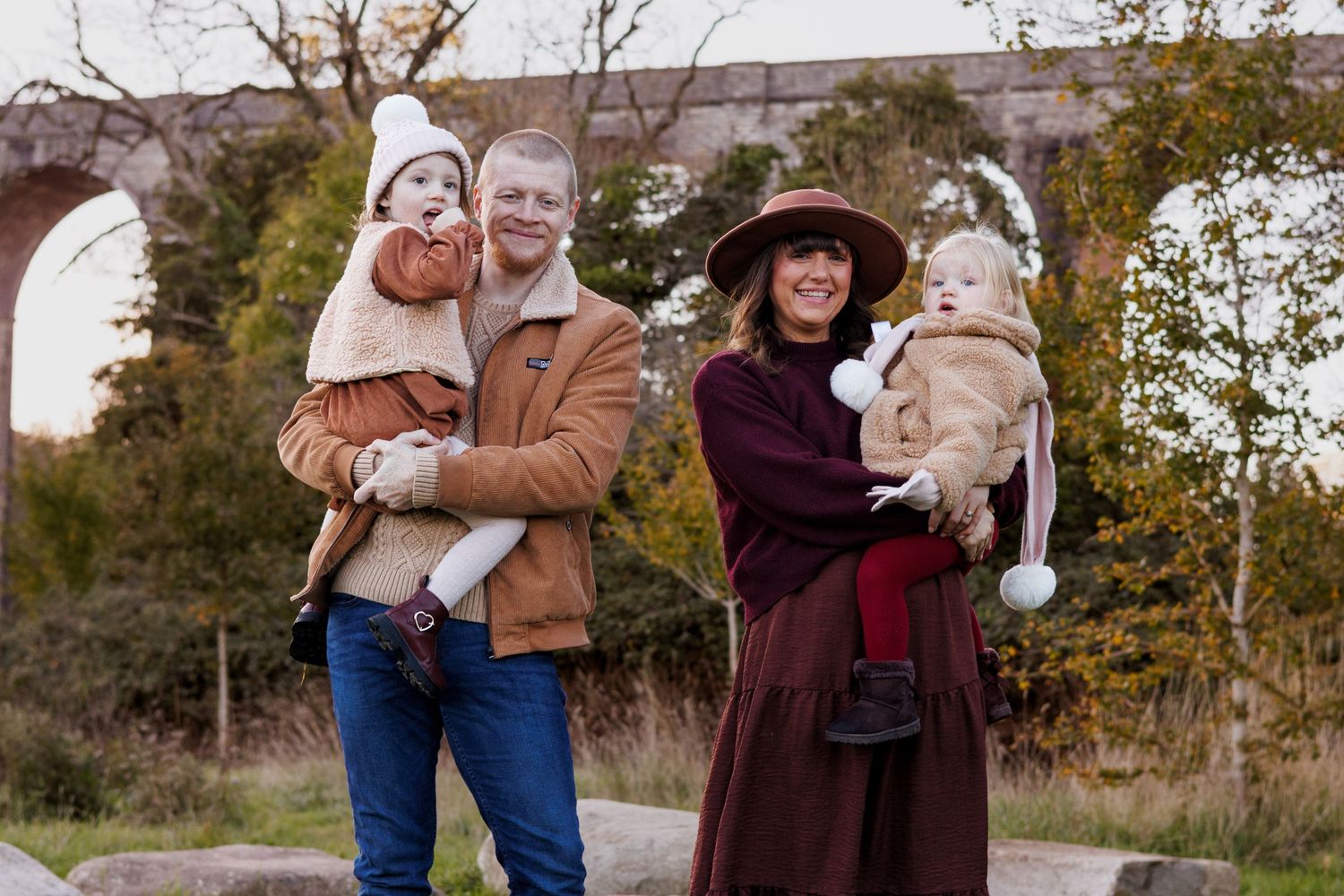 Happy family with two toddlers posing outdoors near stone arched viaduct in autumn foliage.