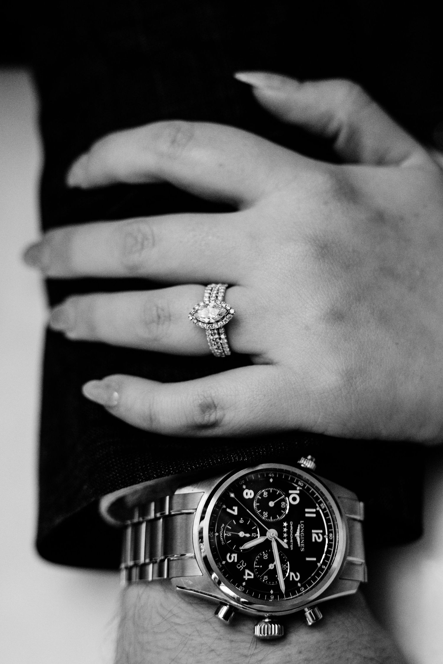 Black and white close-up of a diamond engagement ring and a men's chronograph watch worn by a couple.