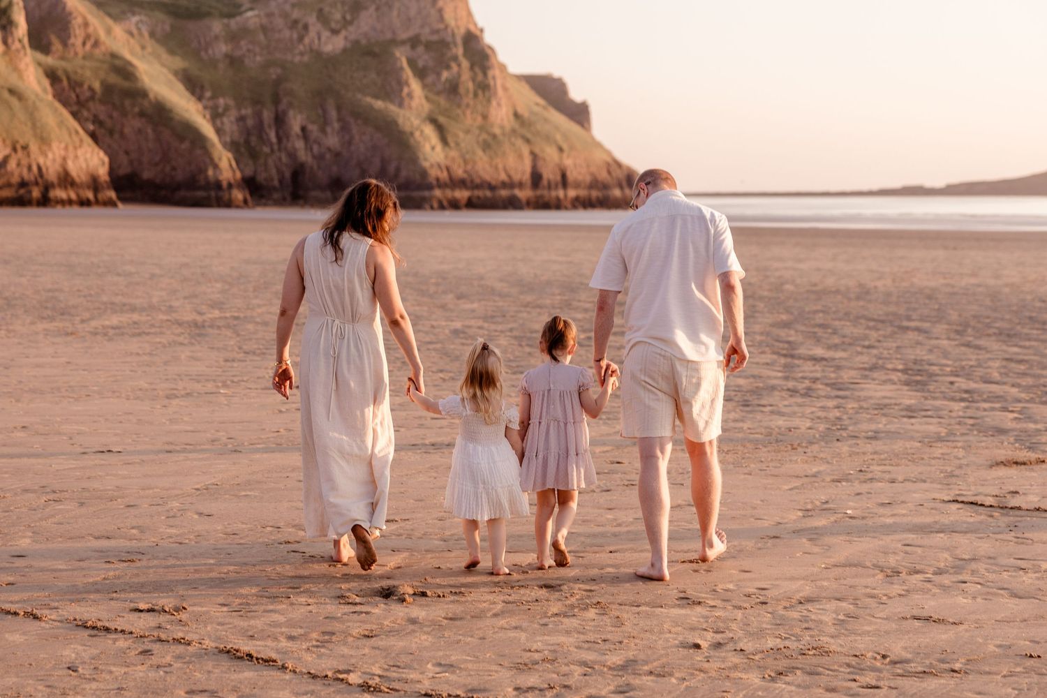 Family of four walking hand-in-hand on a sandy beach at sunset, with cliffs in the background.