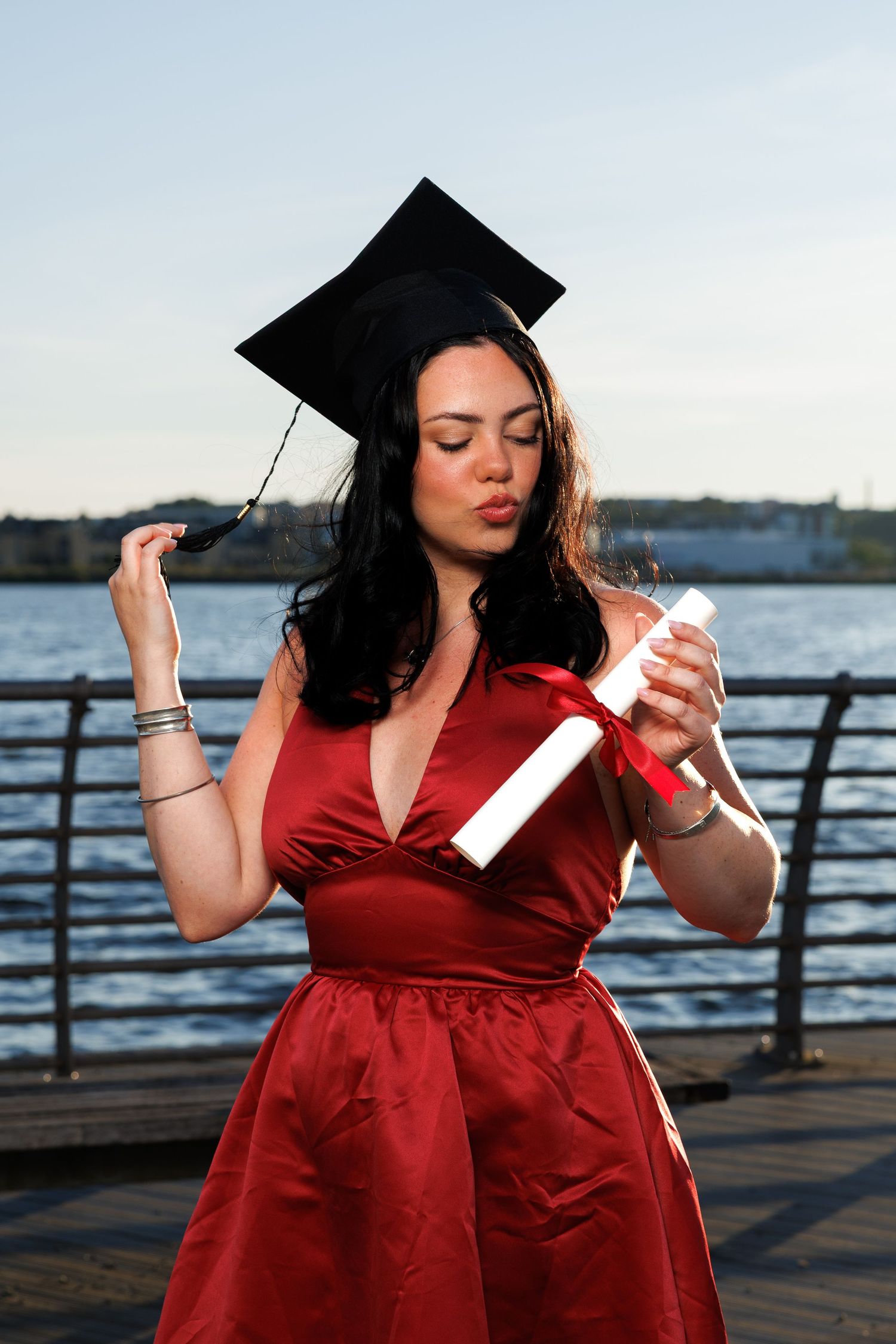 Young woman in red dress and graduation cap holds diploma by waterfront, celebrating academic achievement.