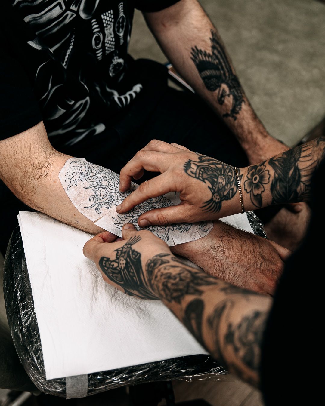 Two heavily tattooed artists collaborating on a hand tattoo design using a stencil.
