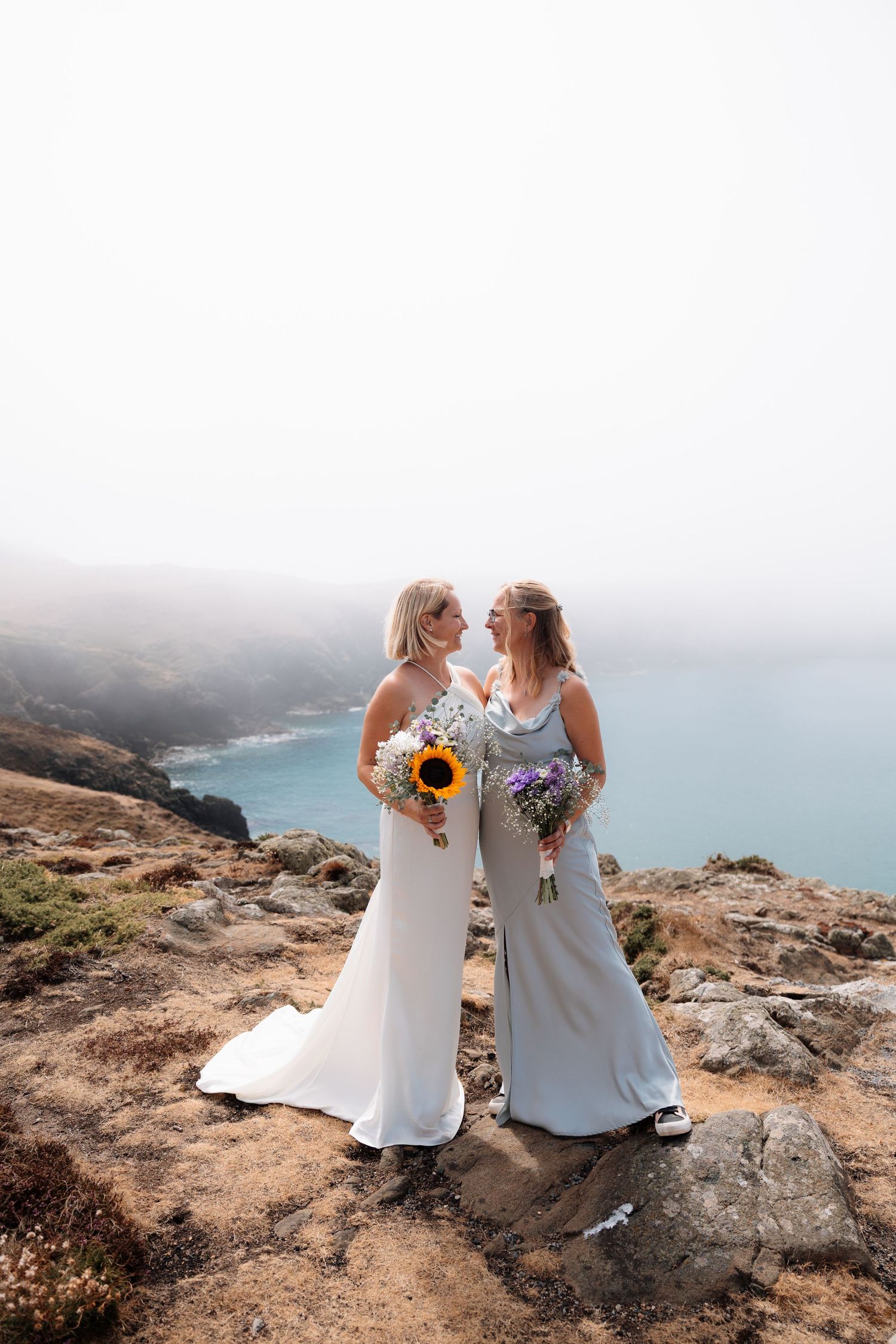 Two brides holding colorful bouquets stand on a coastal cliff overlooking the ocean on their wedding day.