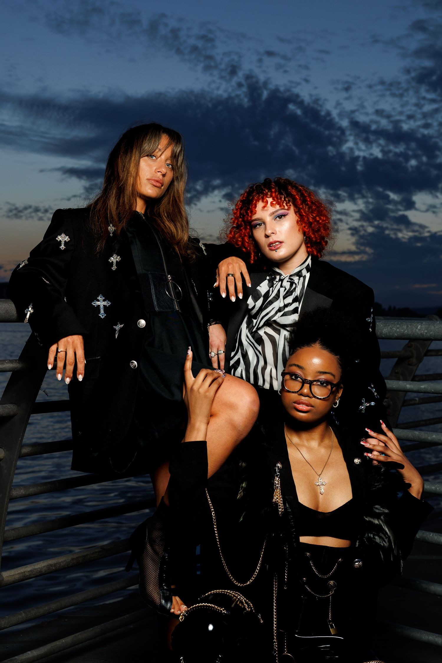 Three stylish women pose on a pier at dusk wearing black embellished jackets with edgy accessories.
