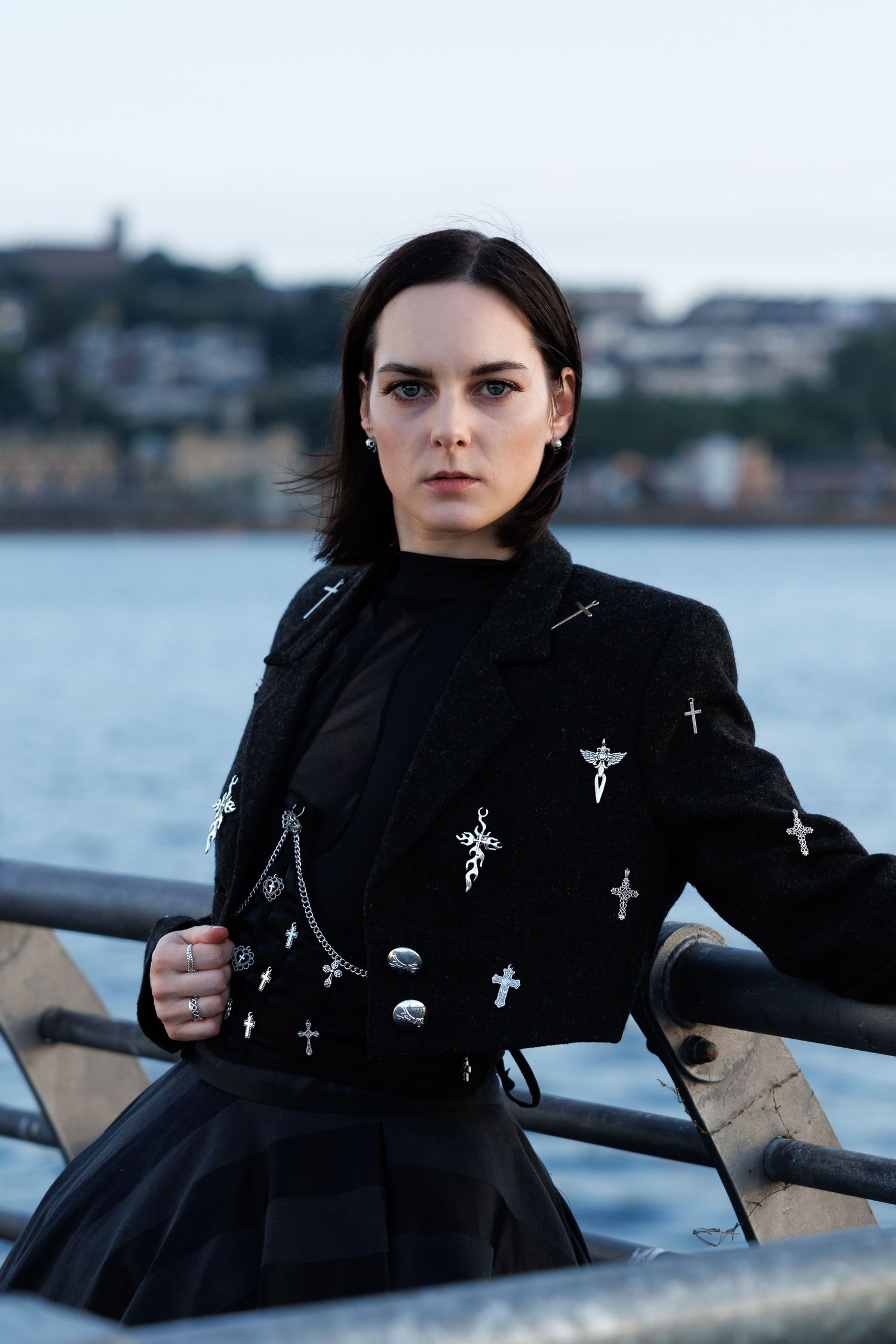 Young woman in black jacket with cross pins leans on metal railing by waterfront at dusk.