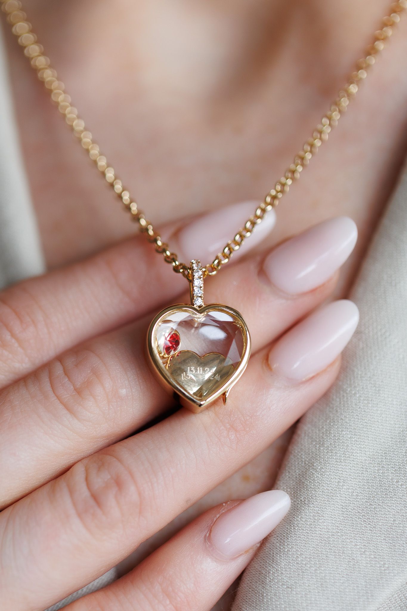 Gold heart locket necklace with diamonds and ruby held by a woman with nude manicured nails.