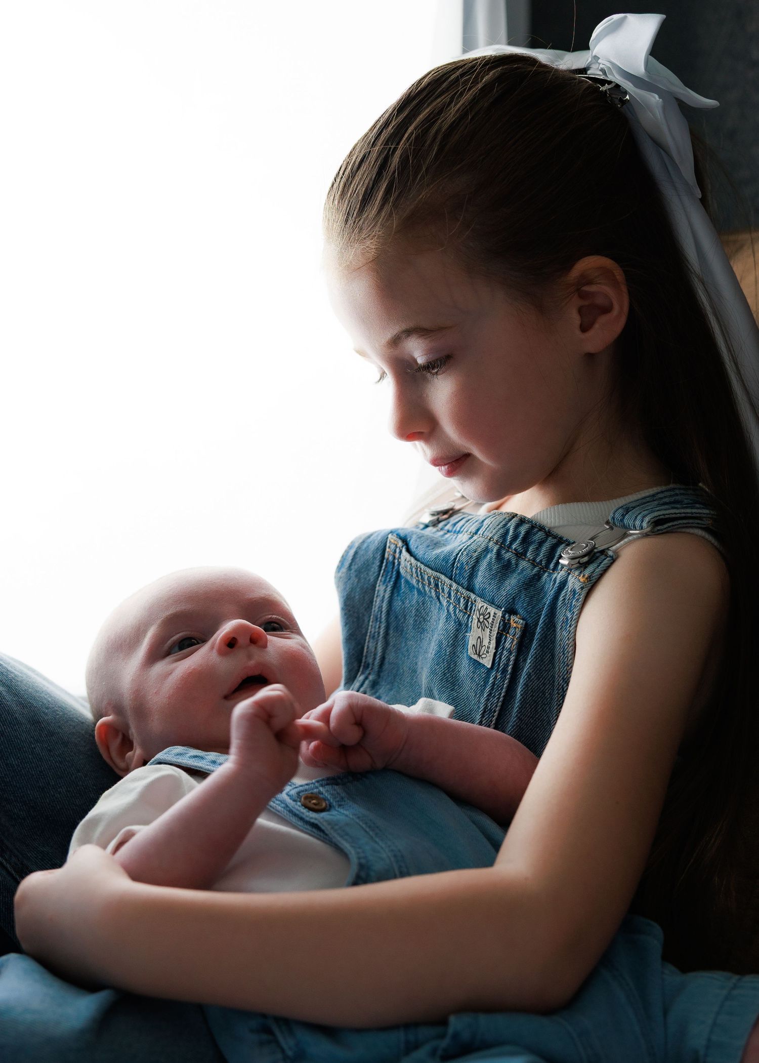 Young girl in denim dress tenderly holds newborn baby, both dressed in blue, in a soft natural light portrait.