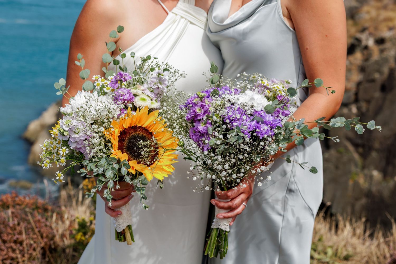 Two bridesmaids in silver gowns holding wildflower bouquets with sunflower and purple blooms by a coastal backdrop.
