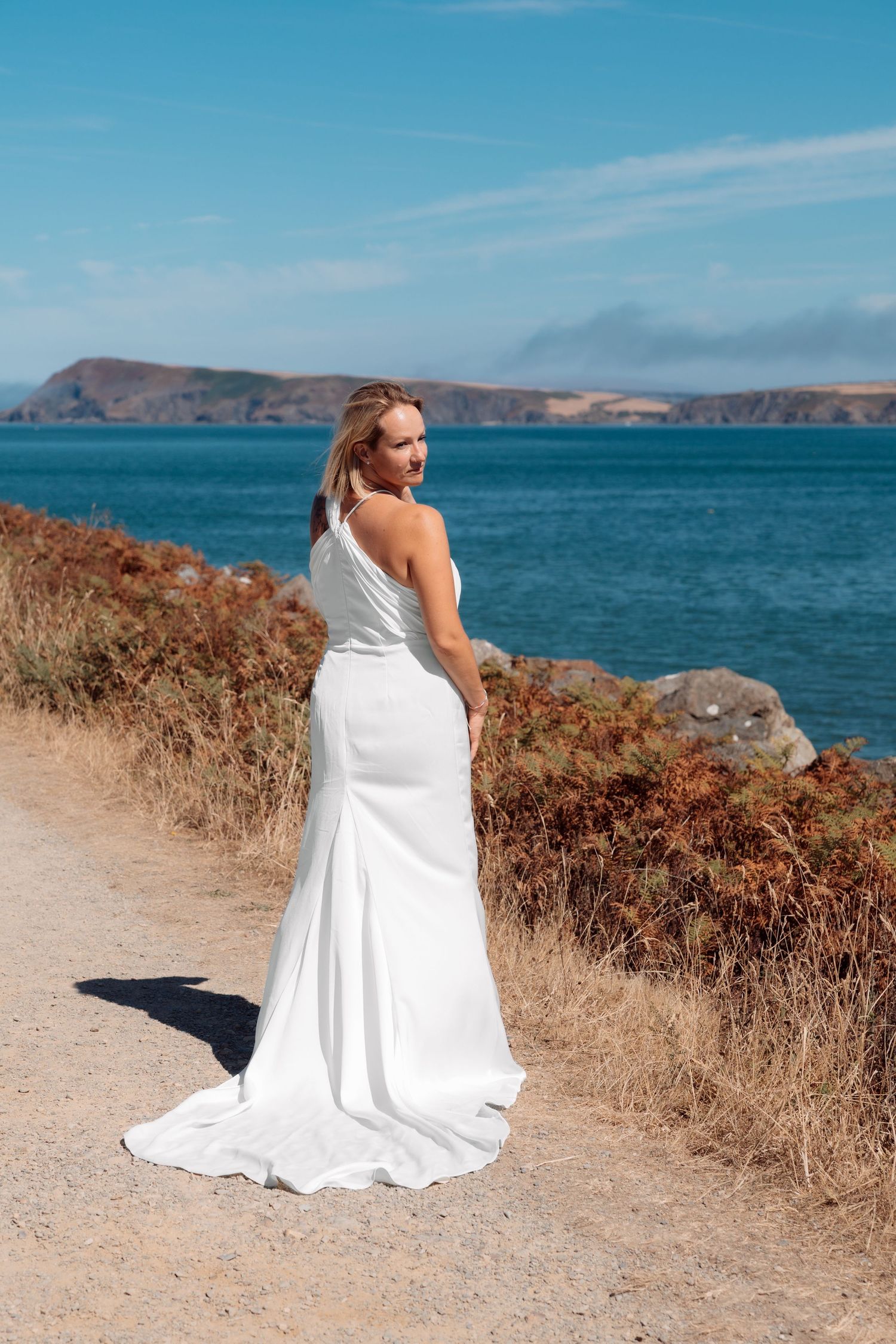 Bride in elegant white gown poses on coastal path with blue ocean and hills in background on sunny day.