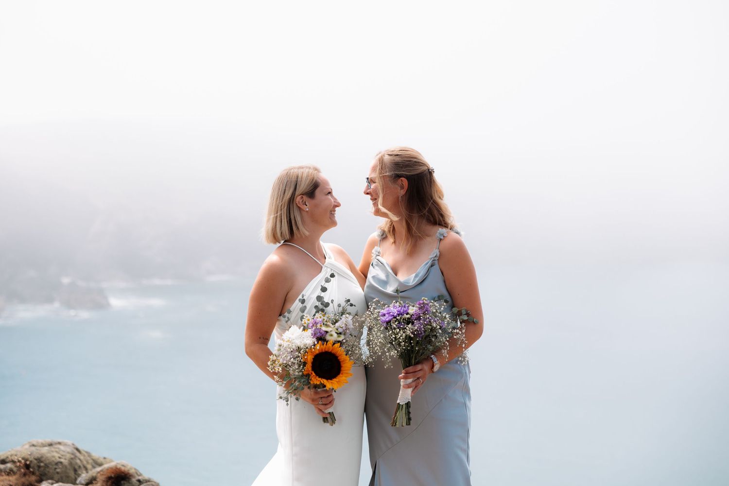 Two brides holding floral bouquets with sunflowers face each other on a coastal cliff with misty ocean views.